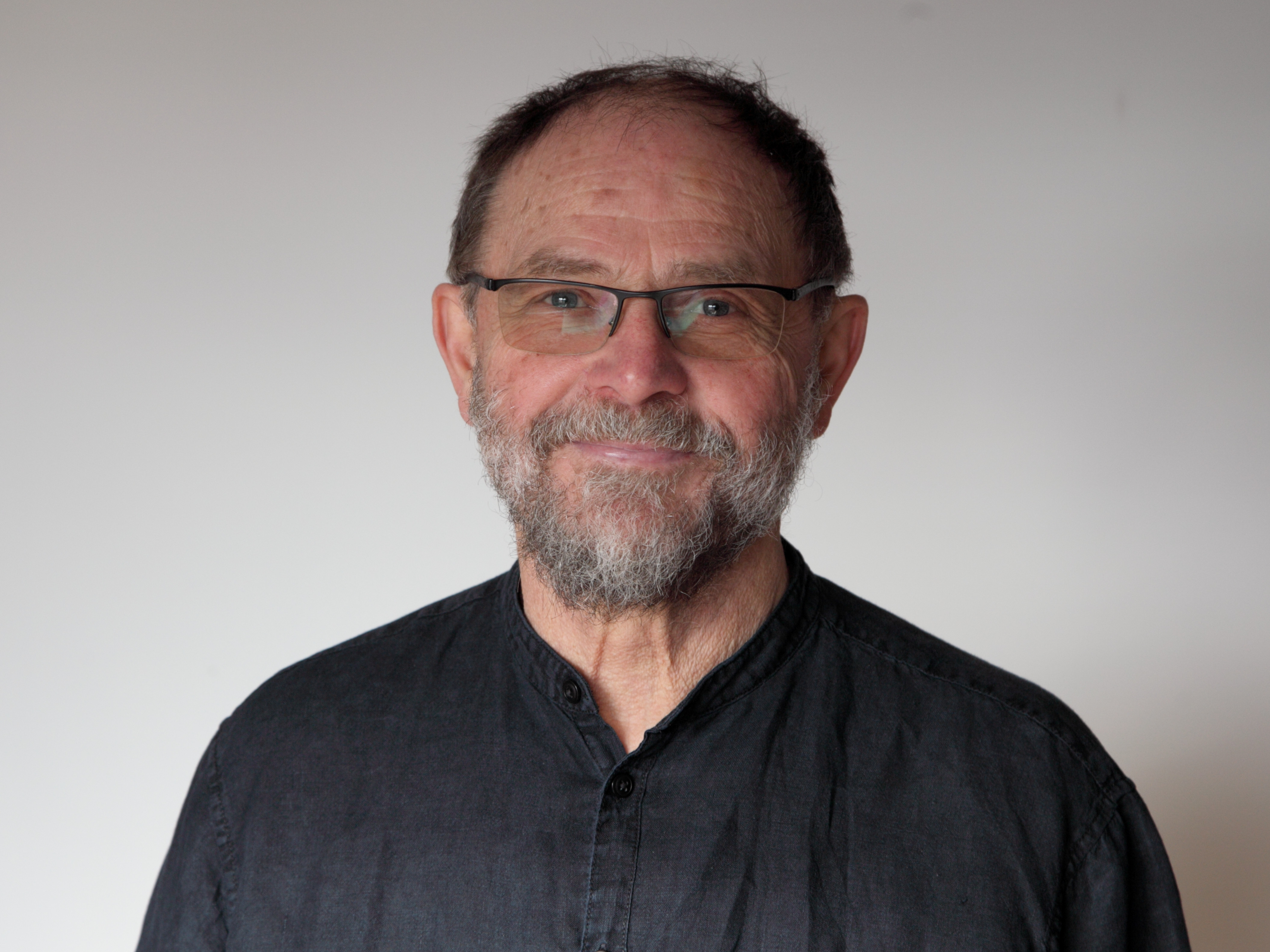 KI generiert: Das Bild zeigt einen Mann mittleren Alters mit Brille und Bart, der freundlich in die Kamera lächelt. Er trägt ein dunkles Hemd vor einem schlichten Hintergrund.