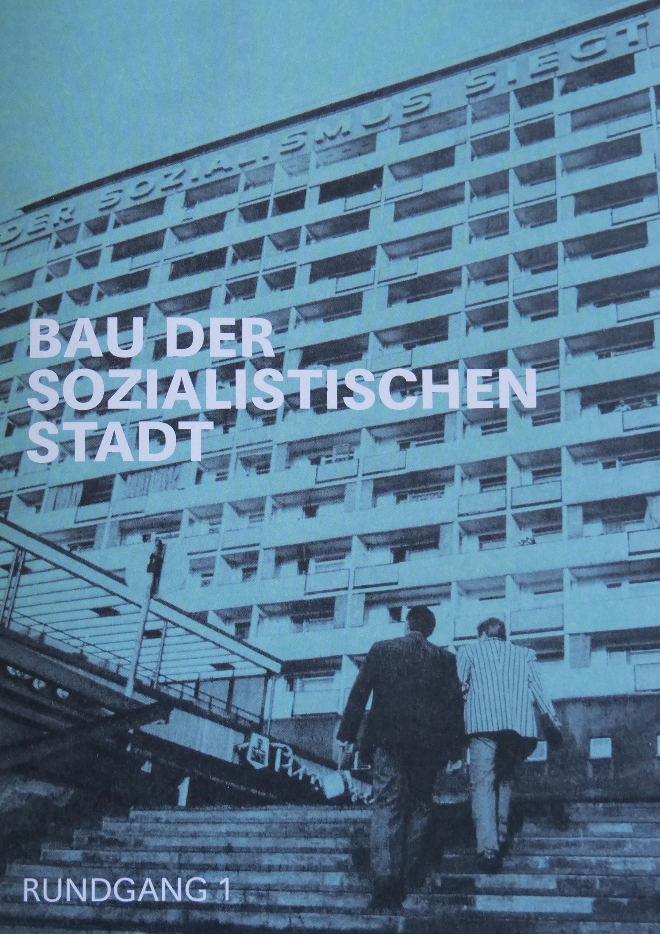 KI generiert: Das Bild zeigt zwei Männer, die eine Treppe zu einem großen, sozialistischen Bauwerk hinaufgehen. Der Text auf dem Bild lautet "Bau der sozialistischen Stadt" und "Rundgang 1".