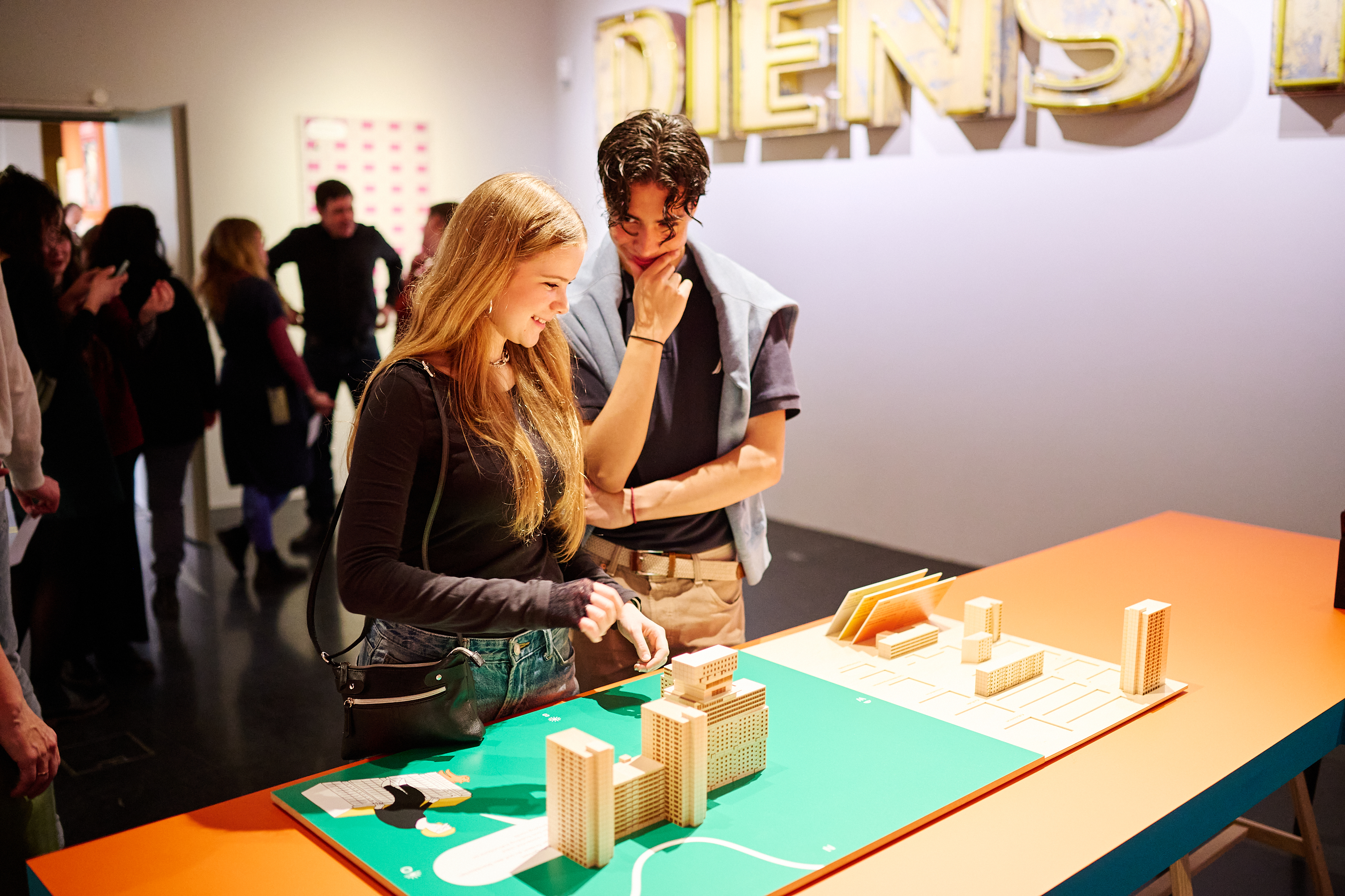 KI generiert: Das Bild zeigt zwei Personen, die ein Modell einer Stadt mit Gebäuden betrachten. Im Hintergrund steht eine Gruppe von Menschen in einem Raum mit der Aufschrift „DIENS“.