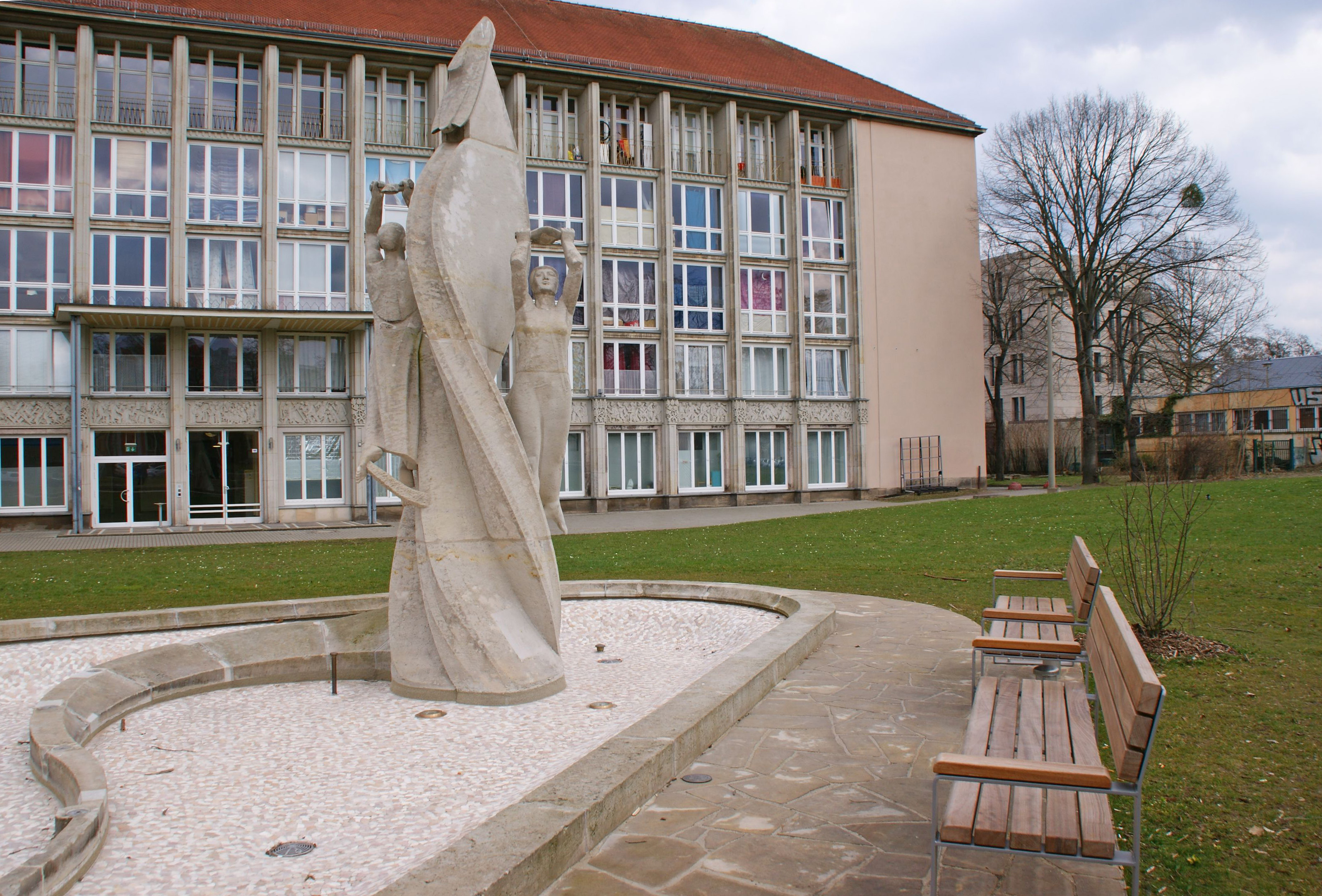 KI generiert: Das Bild zeigt eine moderne Skulpturengruppe aus Stein, die vor einem Gebäude mit mehreren Fenstern und einer Grünfläche aufgestellt ist. Daneben stehen einige Bänke auf einem gepflasterten Weg.