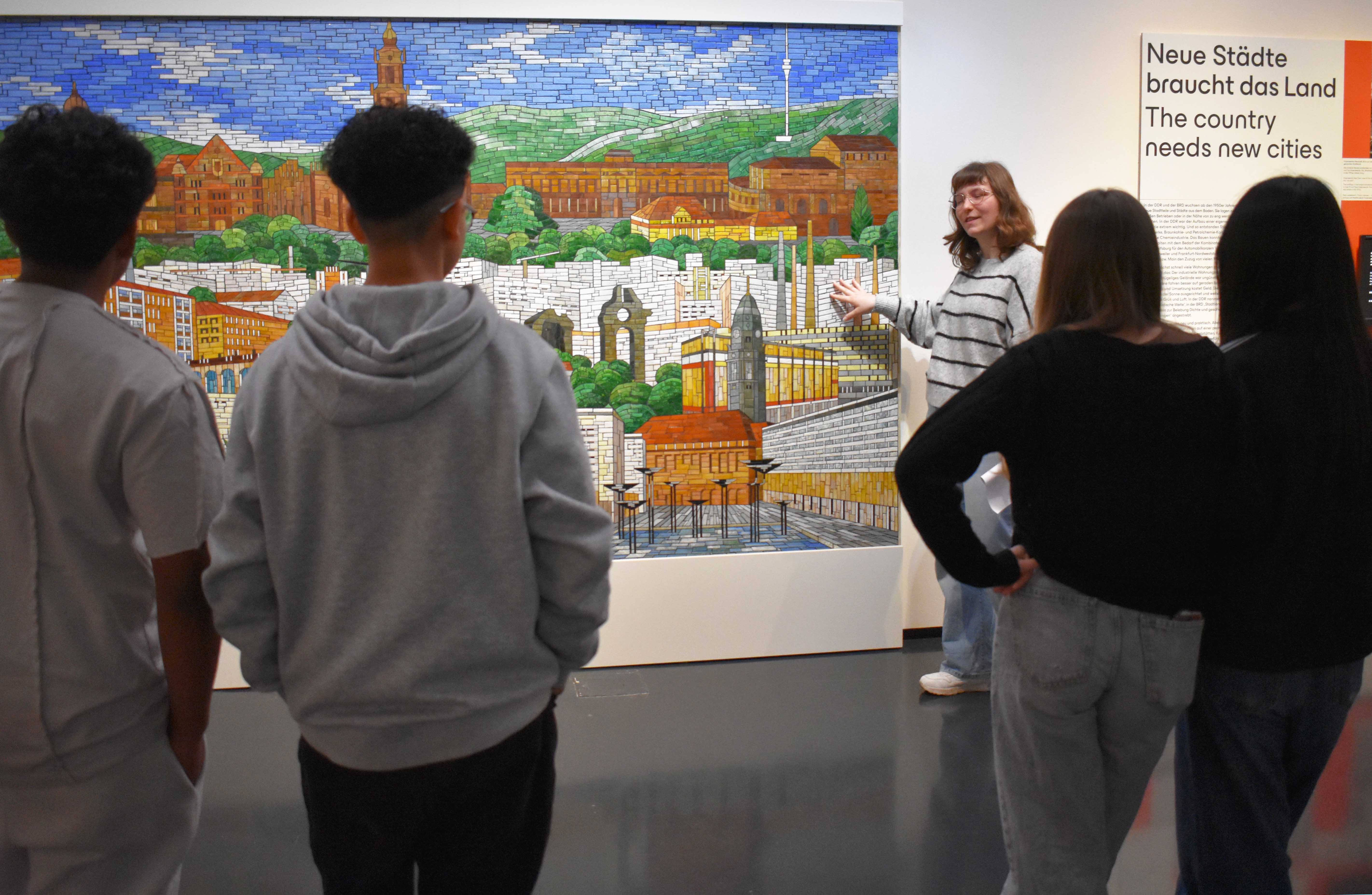 KI generiert: Eine Gruppe von Personen betrachtet ein großes, detailliertes Mosaik, das eine Szenerie zeigt, während eine Führerin darauf hinweist und erklärt. Im Hintergrund ist ein Text auf Deutsch und Englisch zu sehen, der über den Bedarf an neuen Städten spricht.