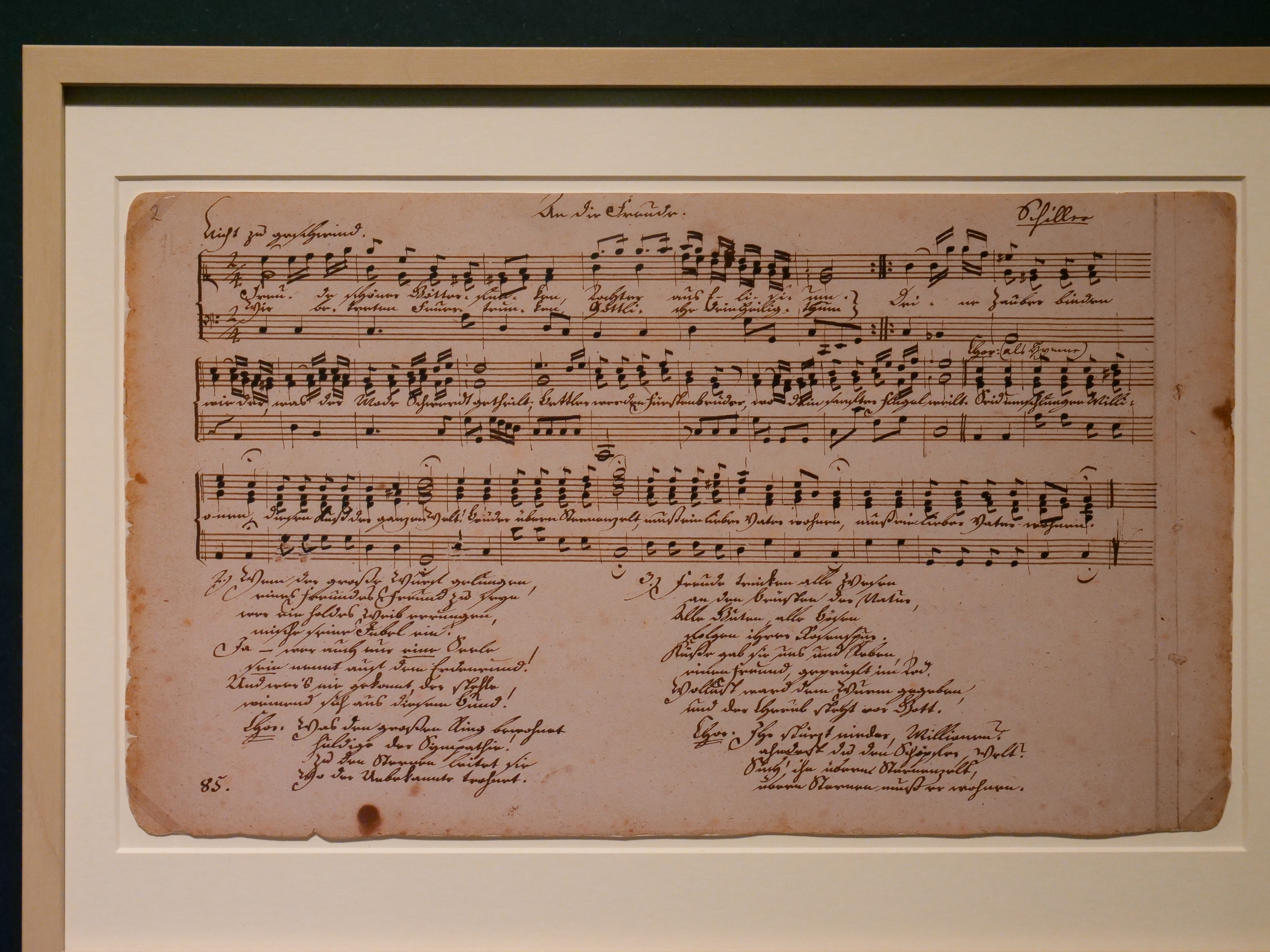 KI generiert: Das Bild zeigt ein altes, handgeschriebenes Musikdokument mit Notenlinien und Texten. Es ist hinter Glas gerahmt und scheint ein historisches Musikstück mit Begleittext zu sein.