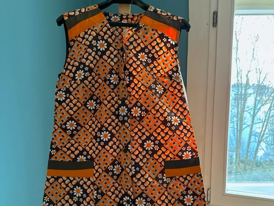 AI generated: The image shows a sleeveless, patterned dress hanging on a hanger. The background is a wall in a light blue colour, next to a window.
