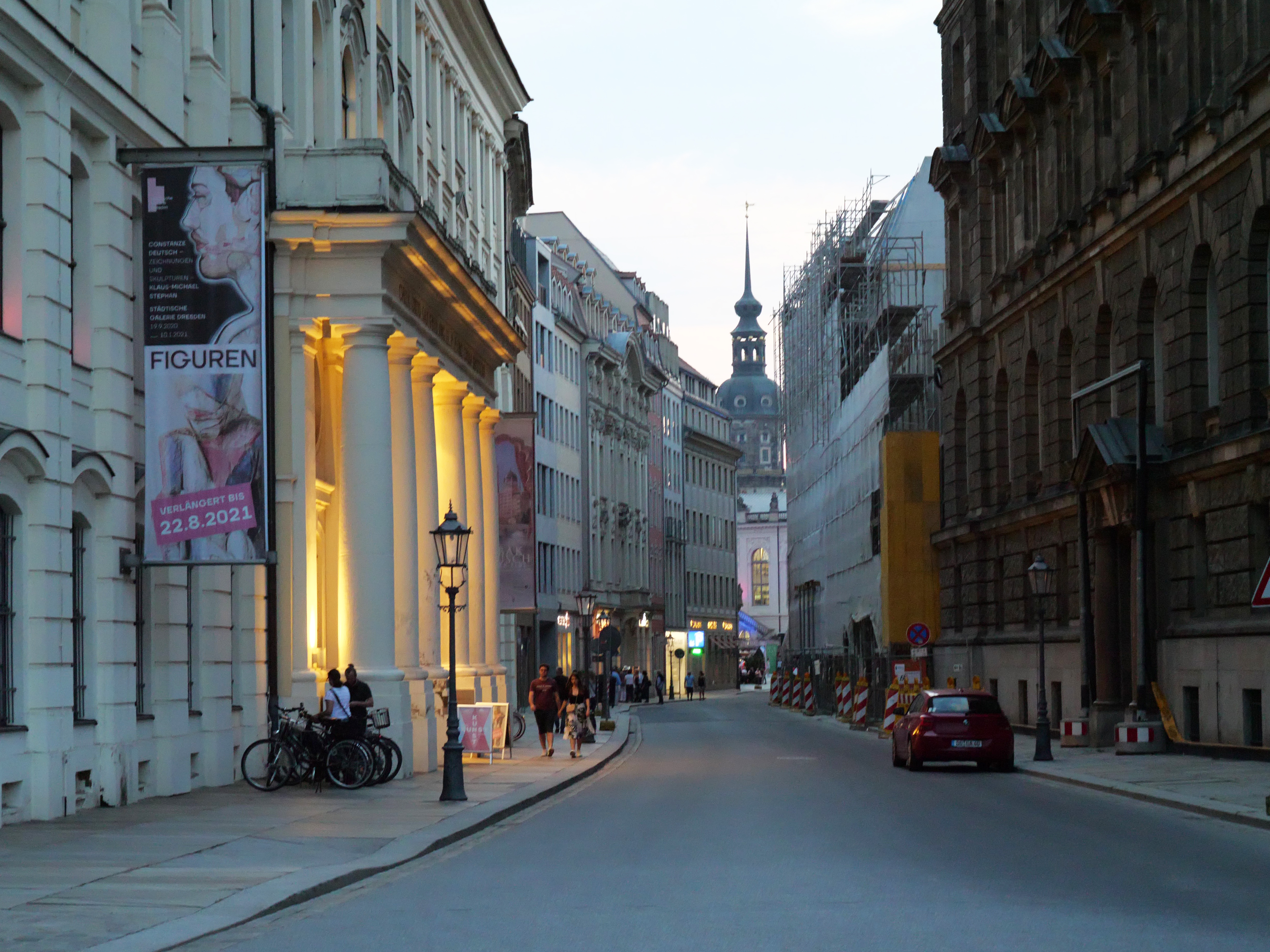 KI generiert: Das Bild zeigt eine schmale Stadtstraße mit historischen Gebäuden auf beiden Seiten, einigen Fahrrädern, Menschen, die spazieren gehen, und einem Auto. Im Hintergrund ist eine Kirche mit einem markanten Turm zu sehen.