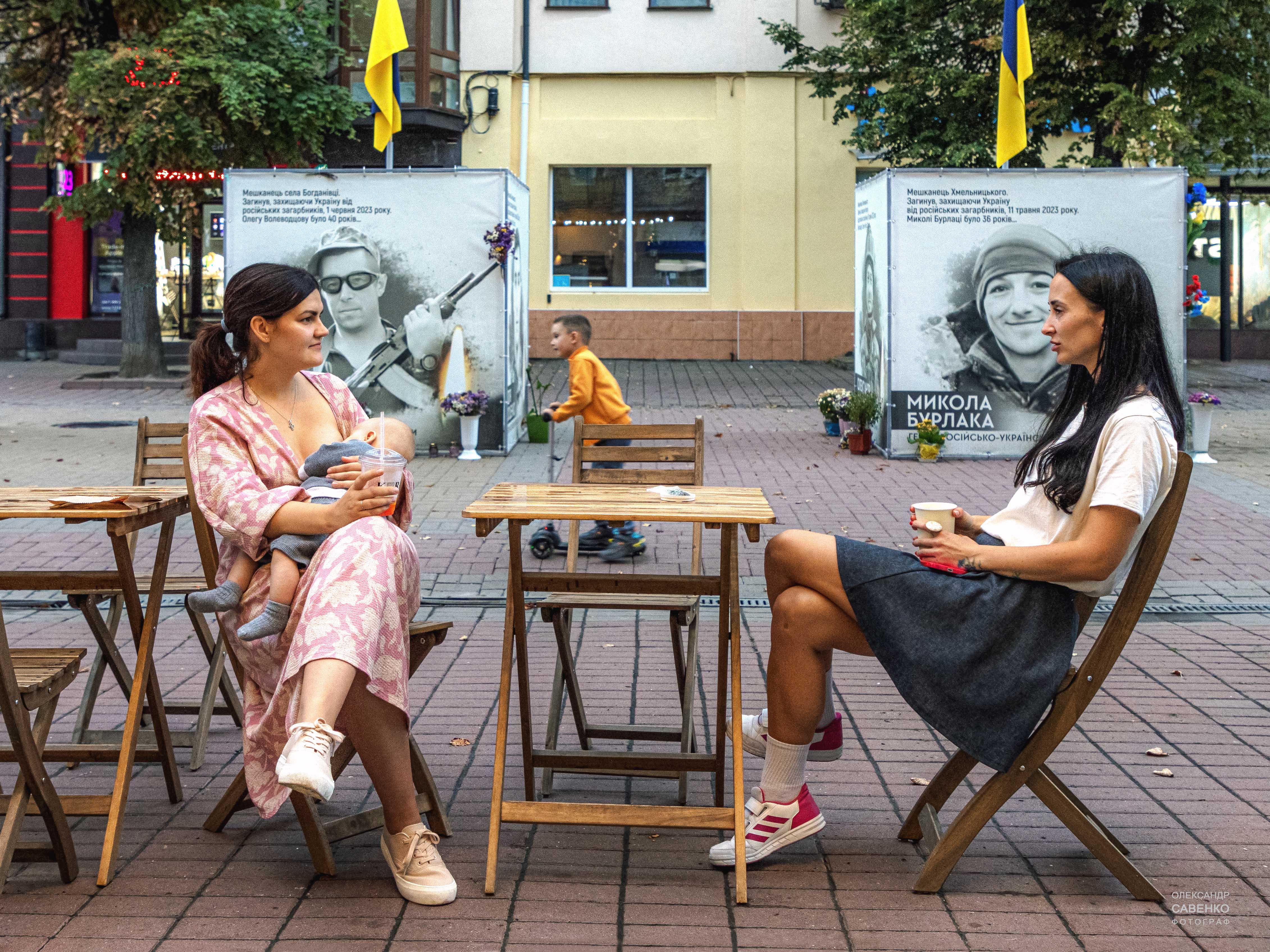 KI generiert: Das Bild zeigt zwei Frauen, die an einem Tisch im Freien sitzen und sich unterhalten, während im Hintergrund ein Kind spielt. Auf Stellwänden sind Porträts und Informationen von Personen abgebildet, flankiert von gelb-blauen Fahnen.