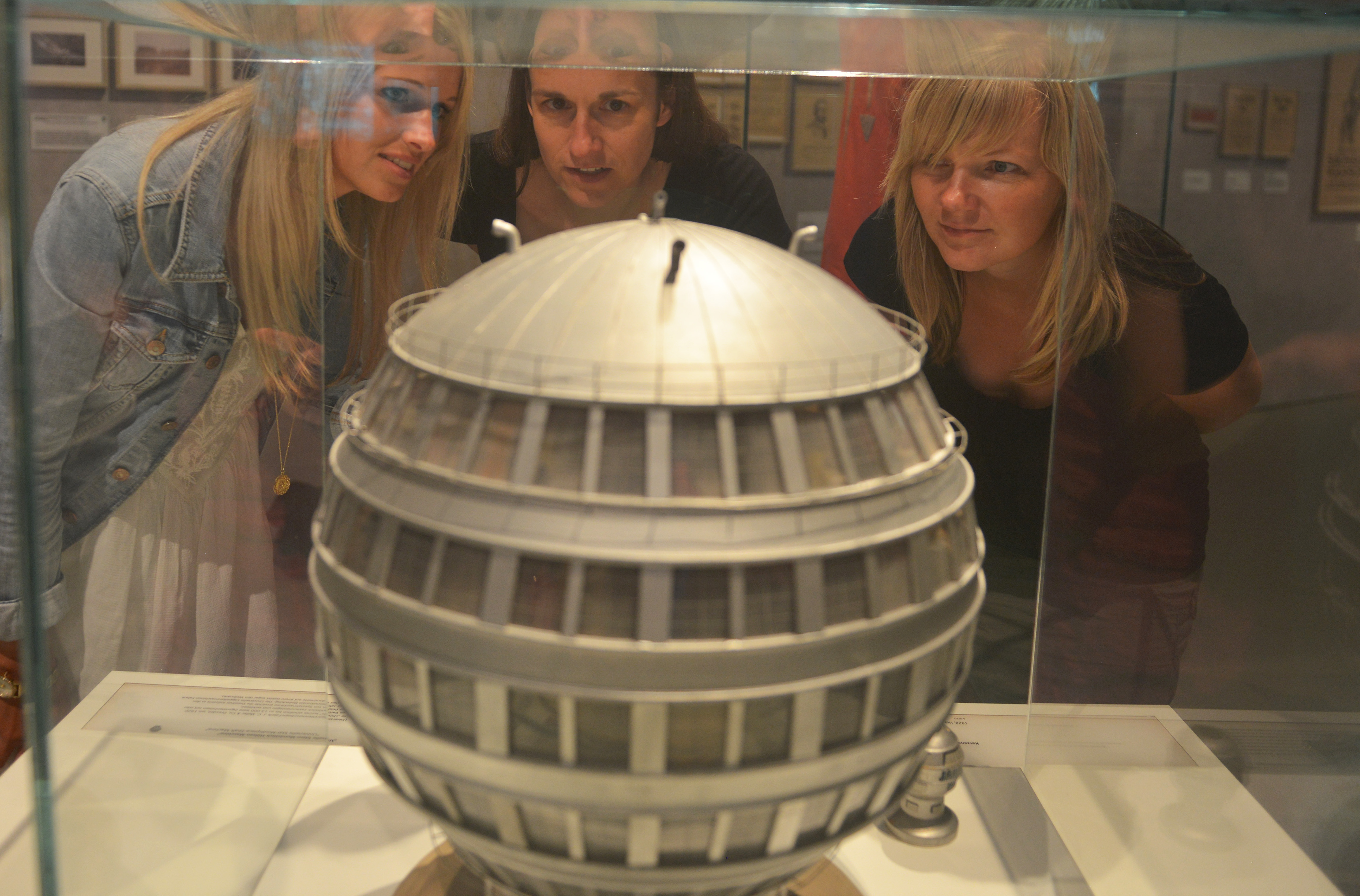 AI-generated: Three people are looking at a model of a spherical building on display in a glass case. The main focus is on the highly detailed spherical model.