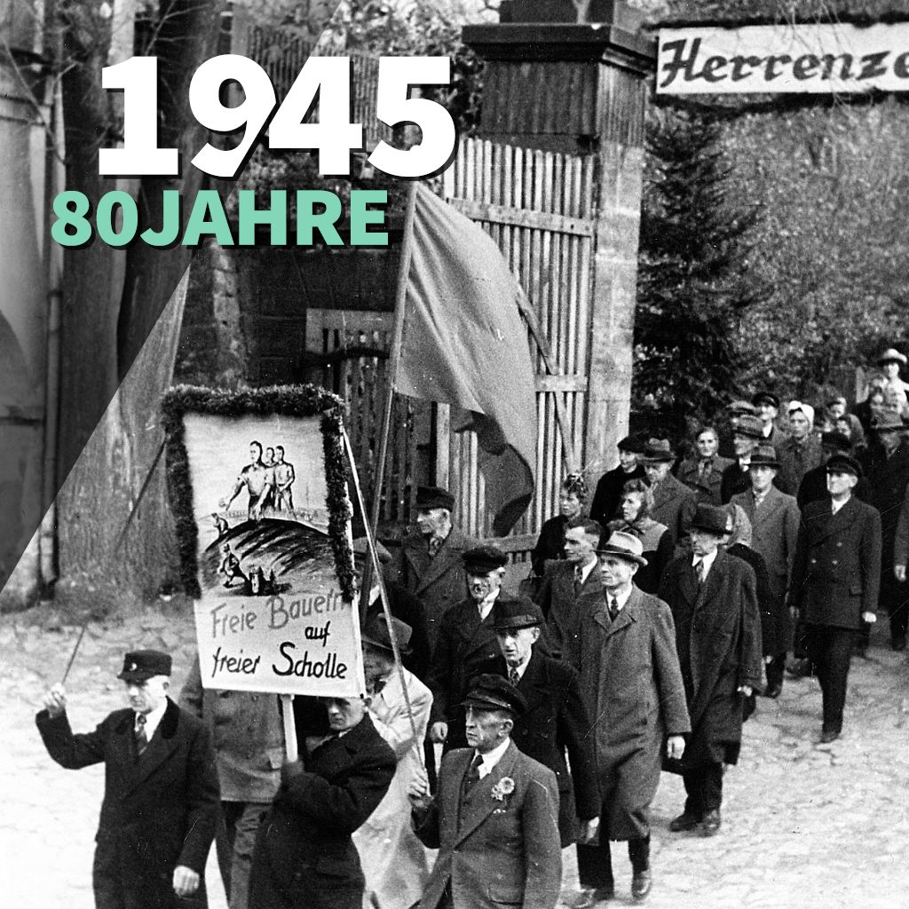 KI generiert: Das Bild zeigt eine historische Demonstration mit einer Gruppe von Menschen, die ein Banner mit der Aufschrift "Freie Bauern auf freier Scholle" und eine rote Fahne tragen. Im Hintergrund ist ein Tor mit der Aufschrift "Herrenz..." zu erkennen, und der Text "1945 80 Jahre" ist im Bild überlagert.