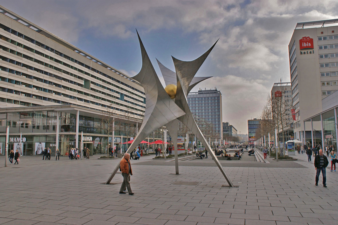 KI generiert: Das Bild zeigt einen offenen Platz in einer Stadt mit modernen Gebäuden und einer auffälligen Skulptur in der Mitte. Menschen spazieren umher, und im Hintergrund sind Geschäfte und ein Hotel zu sehen.
