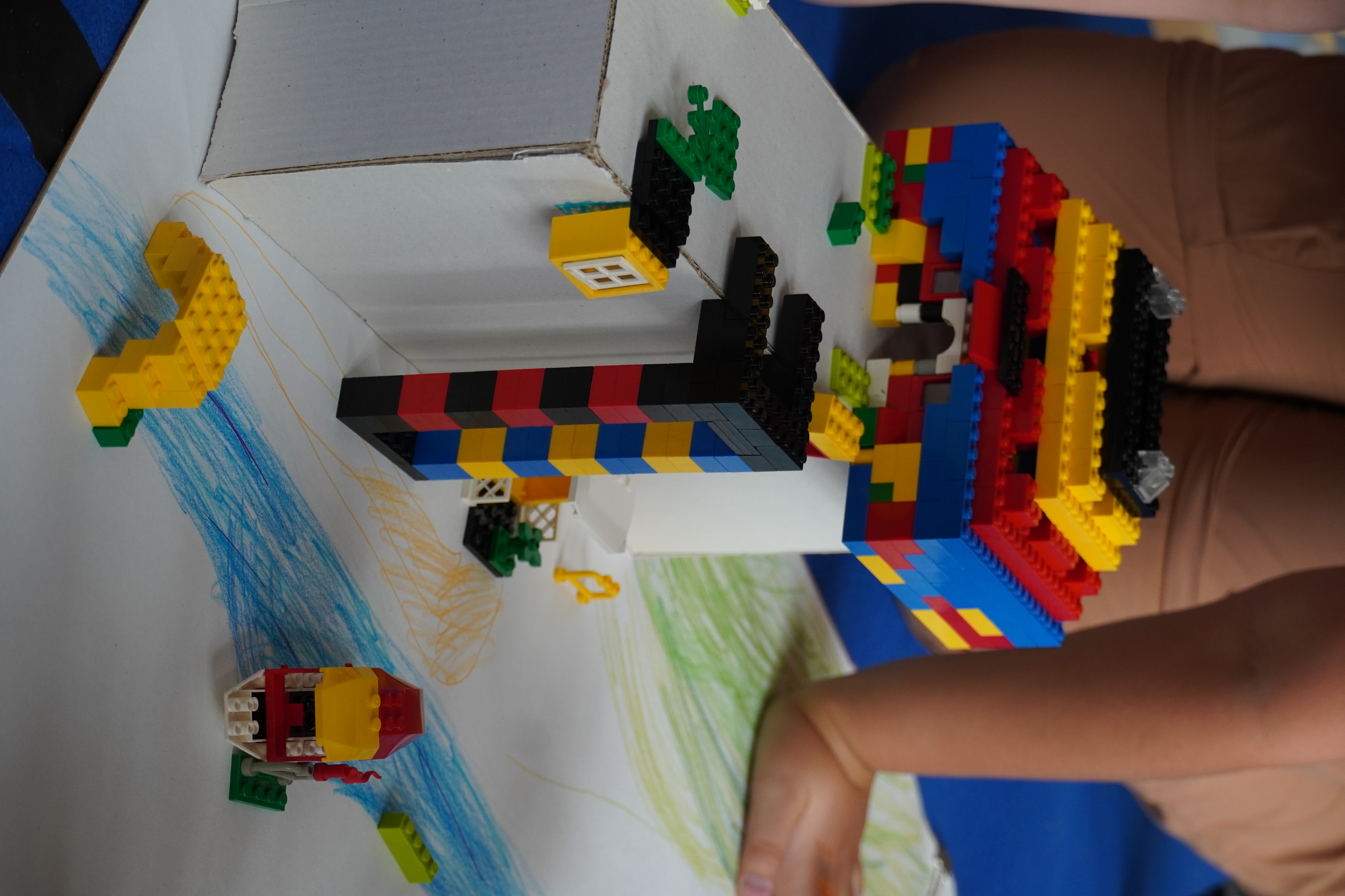 AI-generated: A child is building a structured scene using colourful Lego bricks on a white cardboard box, complemented by a drawn river and green areas. The structure consists of a multicoloured tower and small details such as a yellow arch and a boat.