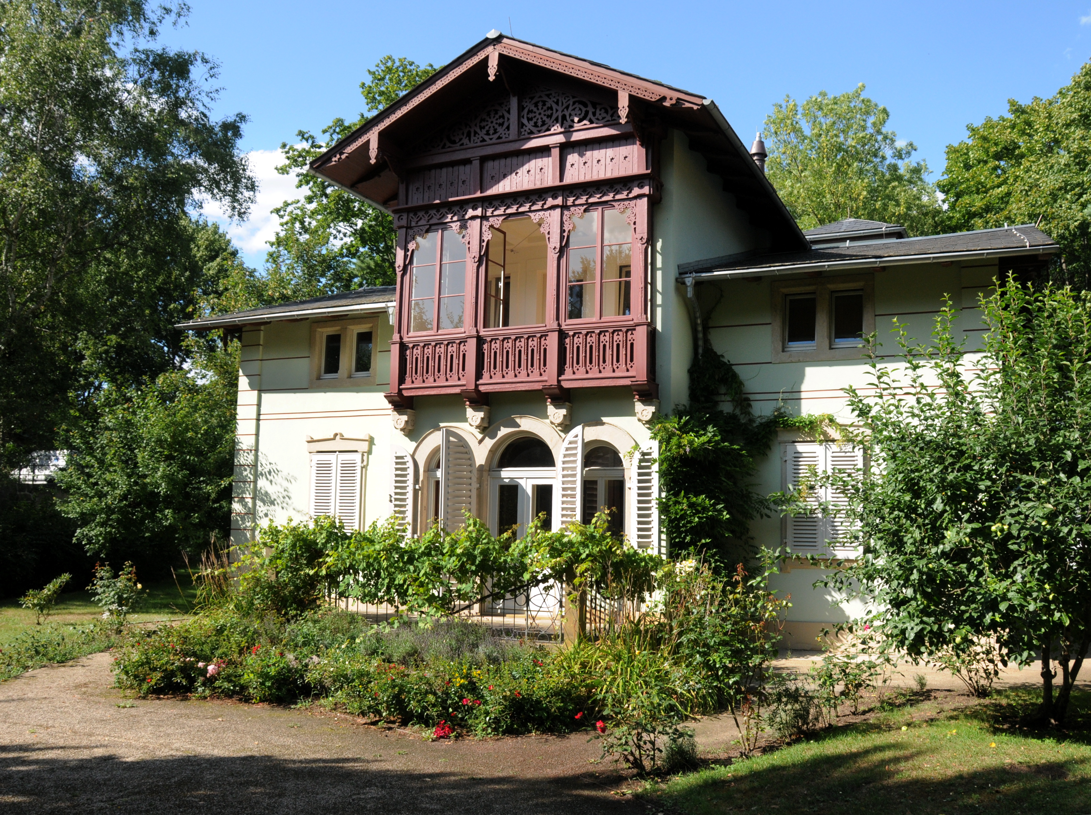 KI generiert: Das Bild zeigt ein charmantes Haus mit einem dekorativen Holzbalkon im obersten Stockwerk, umgeben von einem gepflegten Garten. Die Fassade des Hauses ist von Pflanzen bewachsen, und es steht inmitten einer grünen, baumreichen Umgebung.