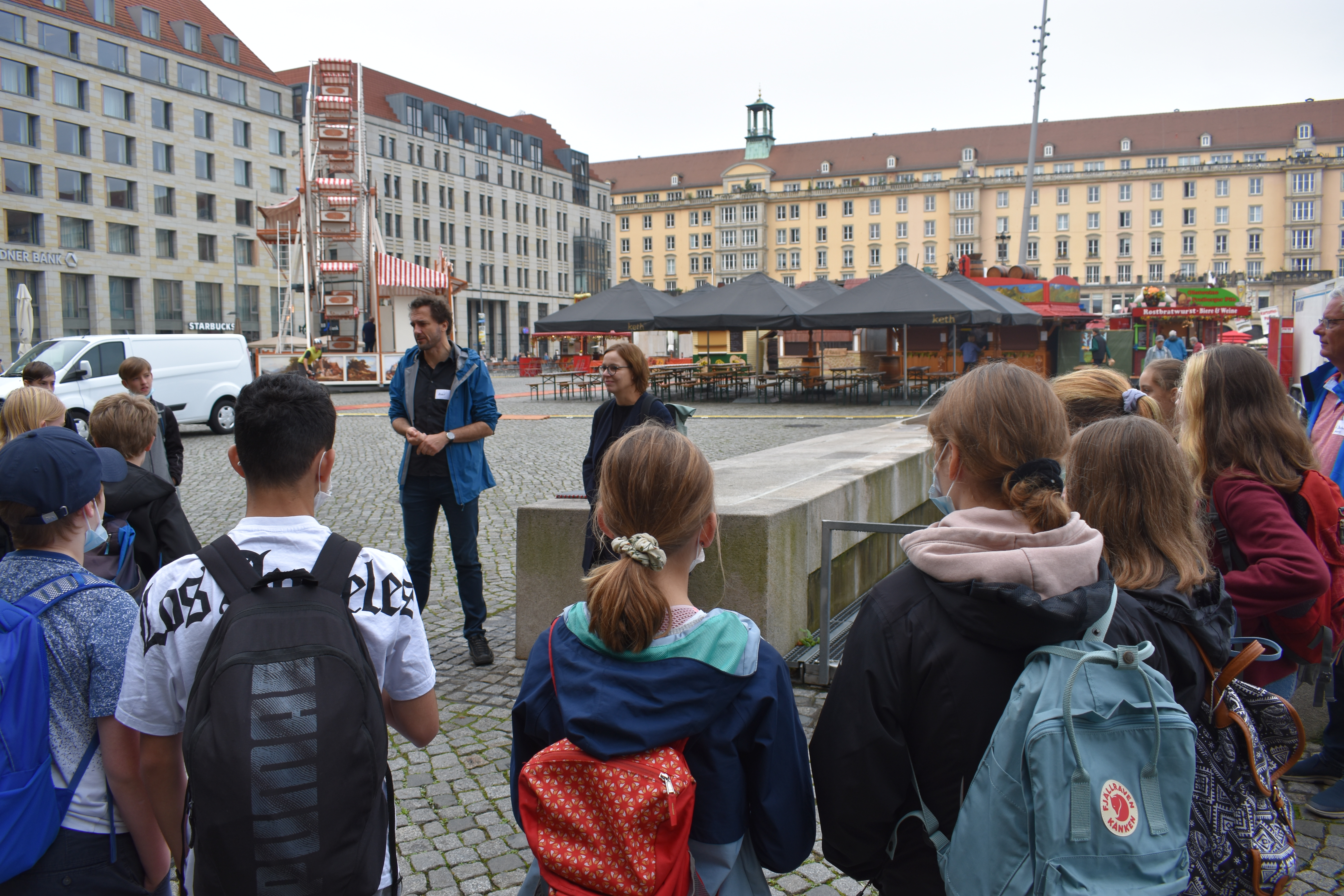 KI generiert: Das Bild zeigt eine Gruppe von Menschen, darunter viele Kinder und Jugendliche, die anscheinend von zwei Erwachsenen auf einem großen, gepflasterten Platz in einer Stadt geführt werden. Im Hintergrund sind Gebäude, Straßencafés und ein kleines Riesenrad zu sehen.