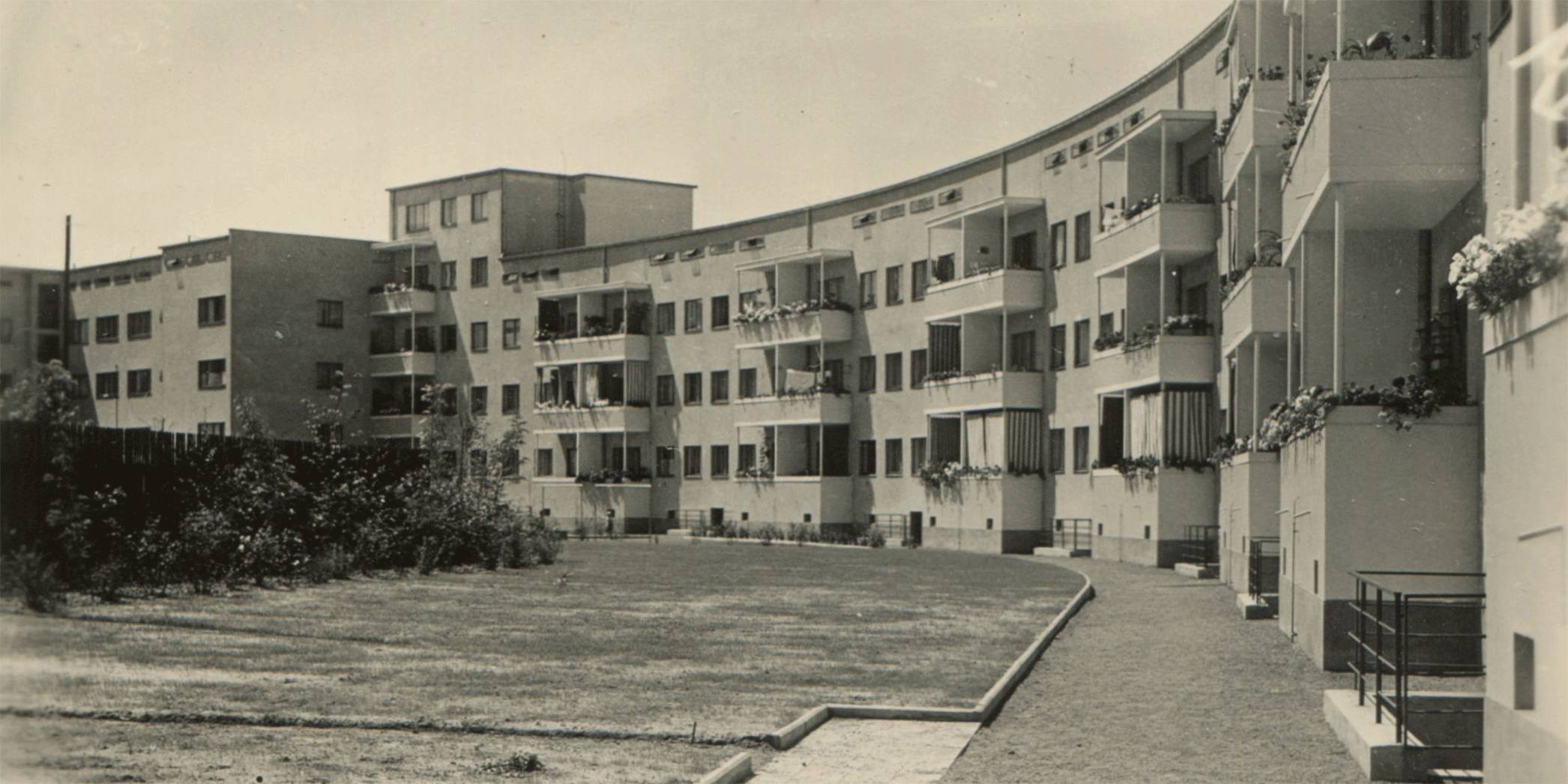 KI generiert: Das Bild zeigt eine geschwungene Reihe von mehrstöckigen Wohngebäuden mit Balkonen, die durch einen gepflegten Garten oder Hof verbunden sind. Der architektonische Stil deutet auf eine moderne, möglicherweise historische Wohnsiedlung hin.