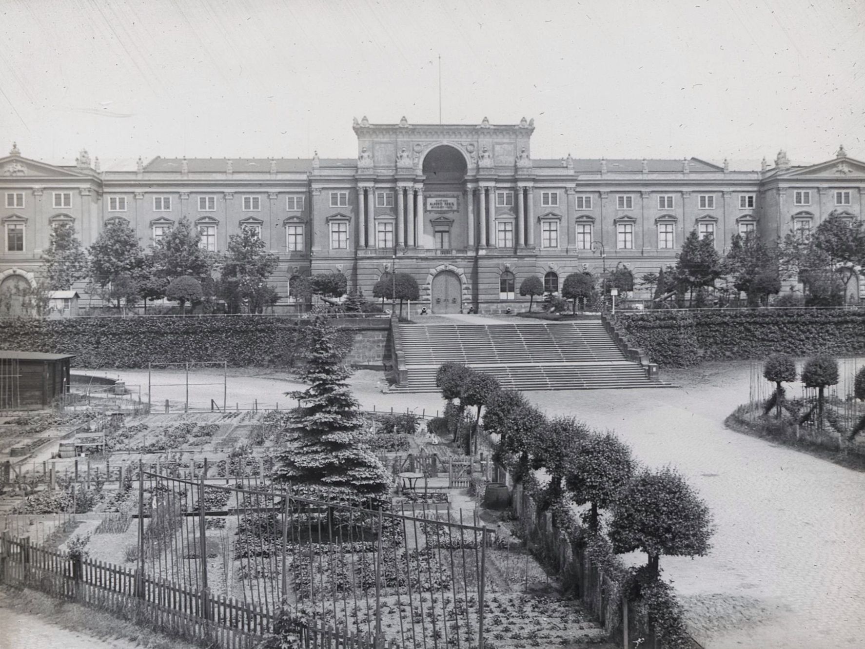 KI generiert: Das Bild zeigt ein großes, historisches Gebäude mit einer prächtigen Fassade und großem Eingangsportal, das über eine breite Treppe erreichbar ist. Vor dem Gebäude befindet sich ein gepflegter Garten mit verschiedenen Pflanzen und Bäumen.