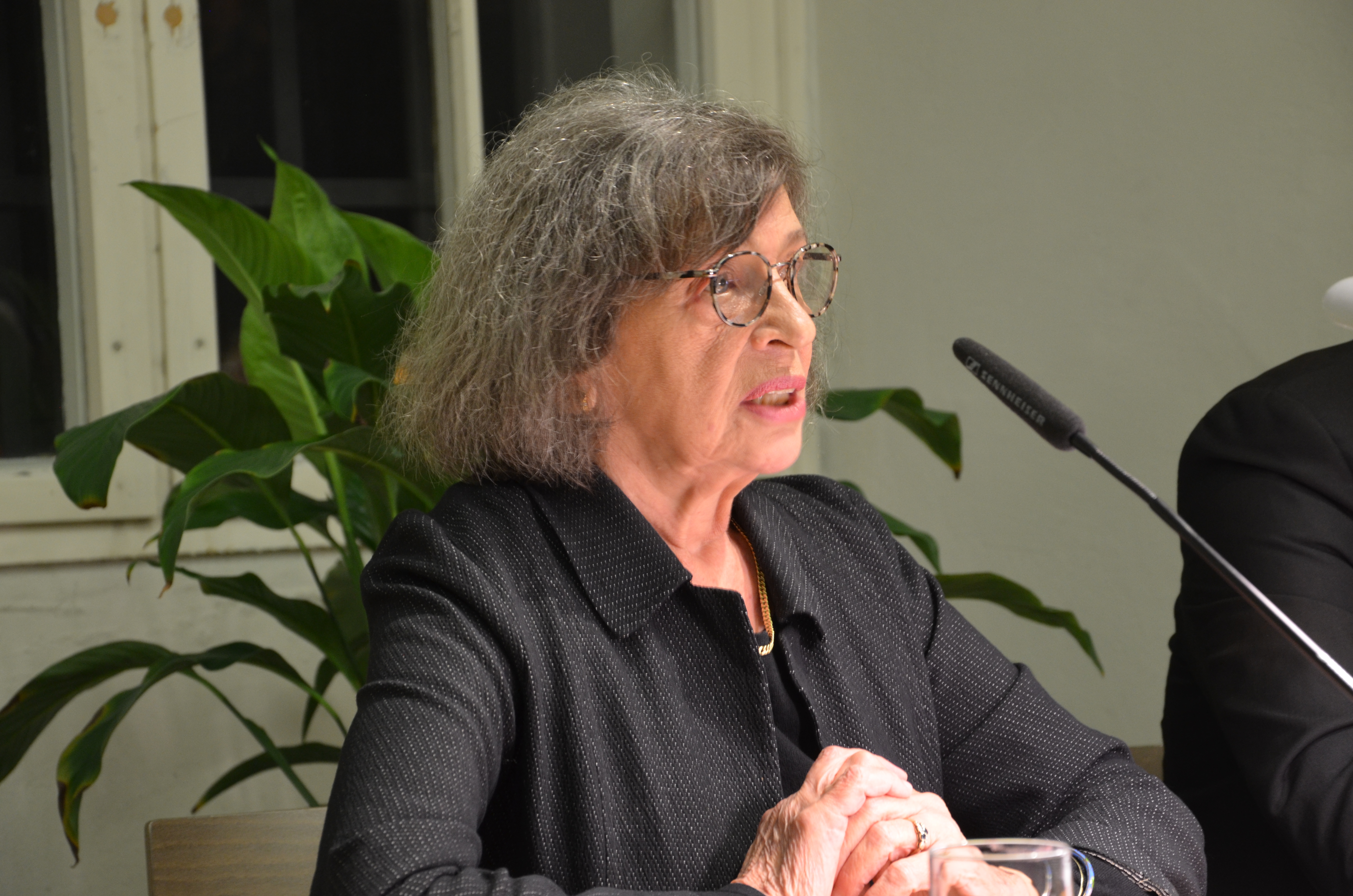 KI generiert: Das Bild zeigt eine ältere Frau mit grauem Haar und Brille, die an einem Tisch sitzt und offenbar an einer Veranstaltung oder Diskussion teilnimmt. Sie spricht in ein Mikrofon, während eine Pflanze im Hintergrund zu sehen ist.