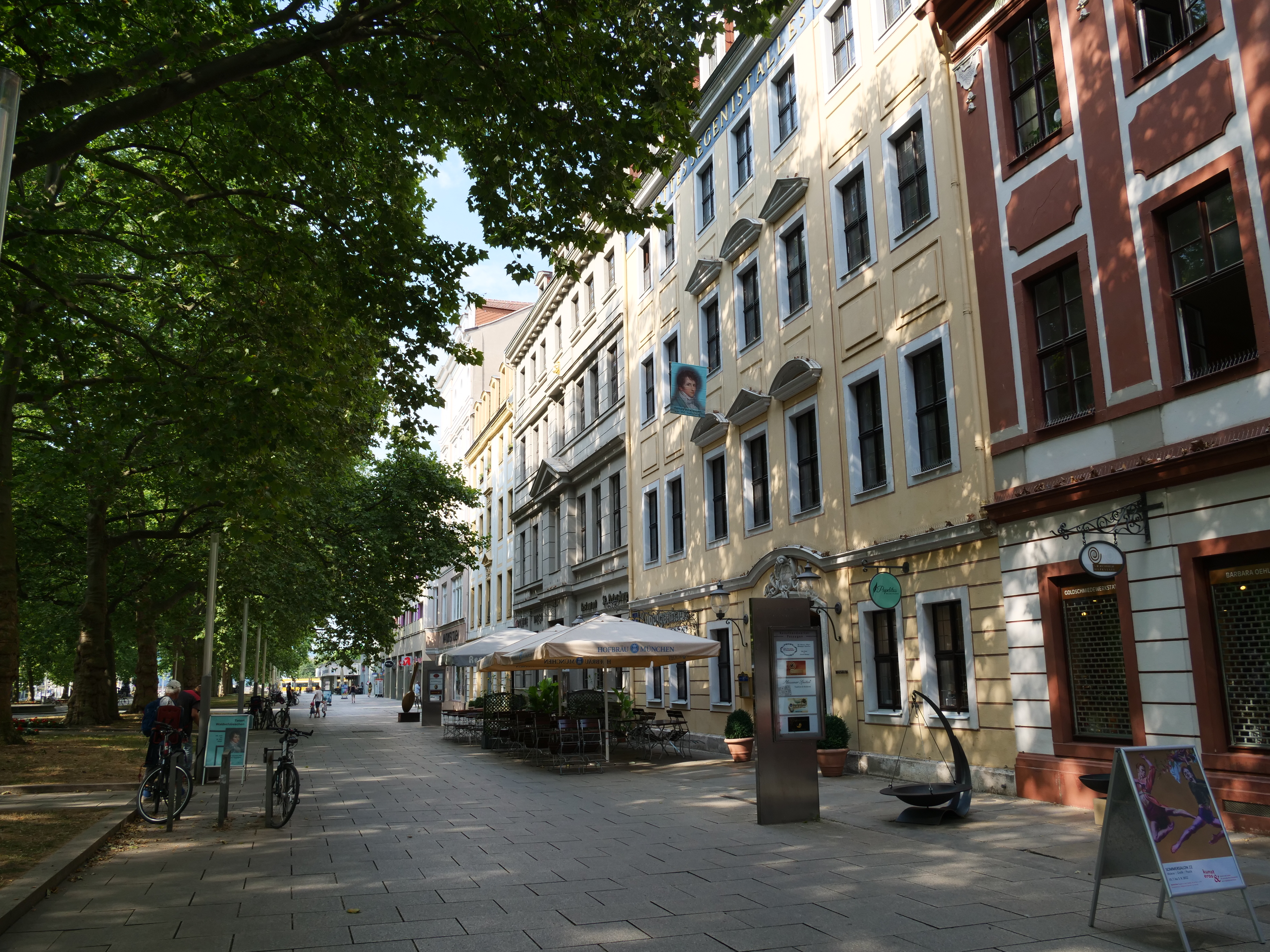 KI generiert: Das Bild zeigt eine belebte Straße mit Cafés und Geschäften, flankiert von mehrstöckigen Gebäuden und einem grünen Park auf der linken Seite. Einige Fahrräder und ein Fußgänger sind ebenfalls zu sehen.