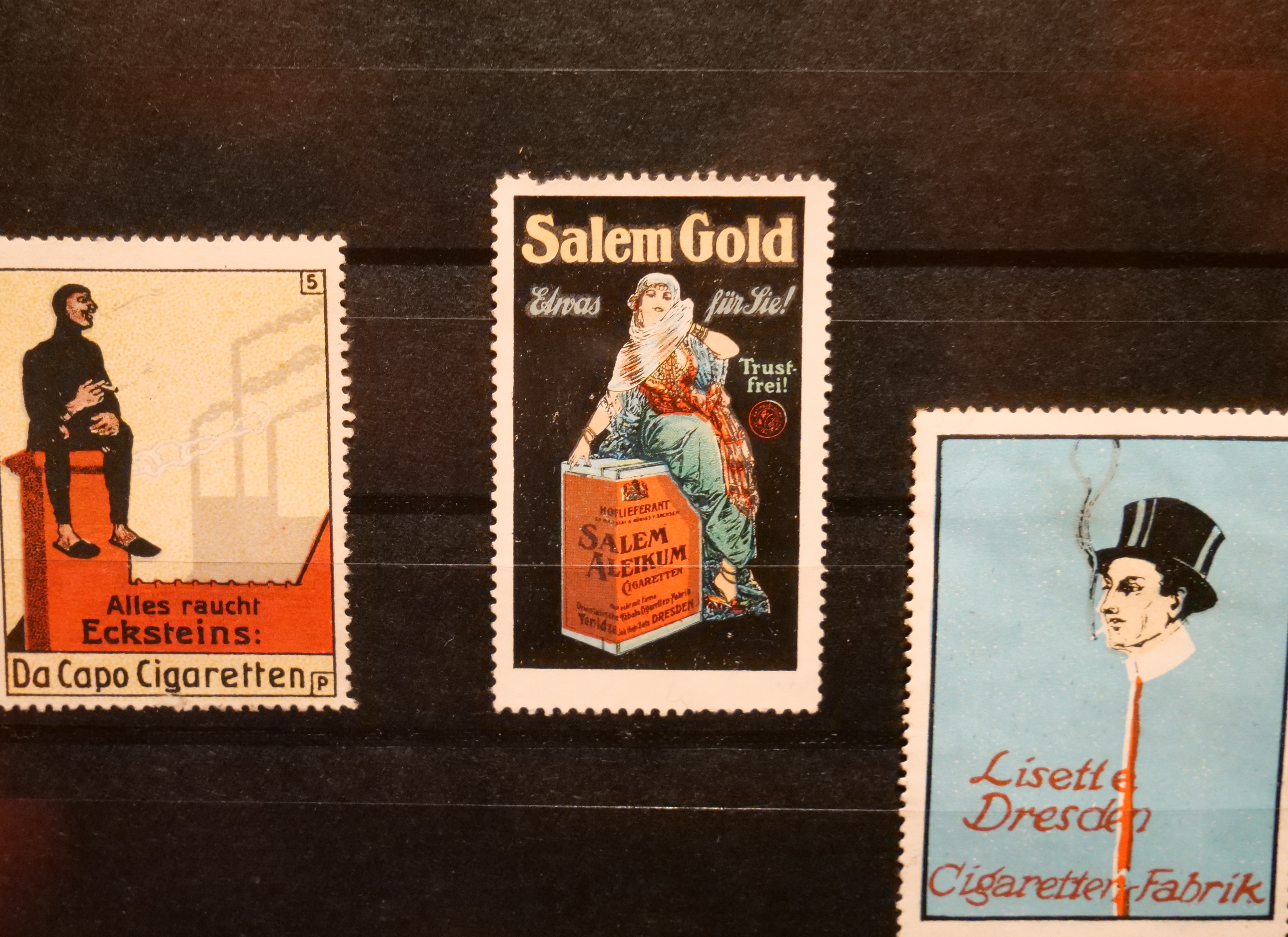 KI generiert: Das Bild zeigt drei historische Zigarettenwerbungen in Form von Sammelbildern. Jede Werbung präsentiert eine andere Zigarettenmarke mit einzigartigen Grafiken und Schriftzügen.