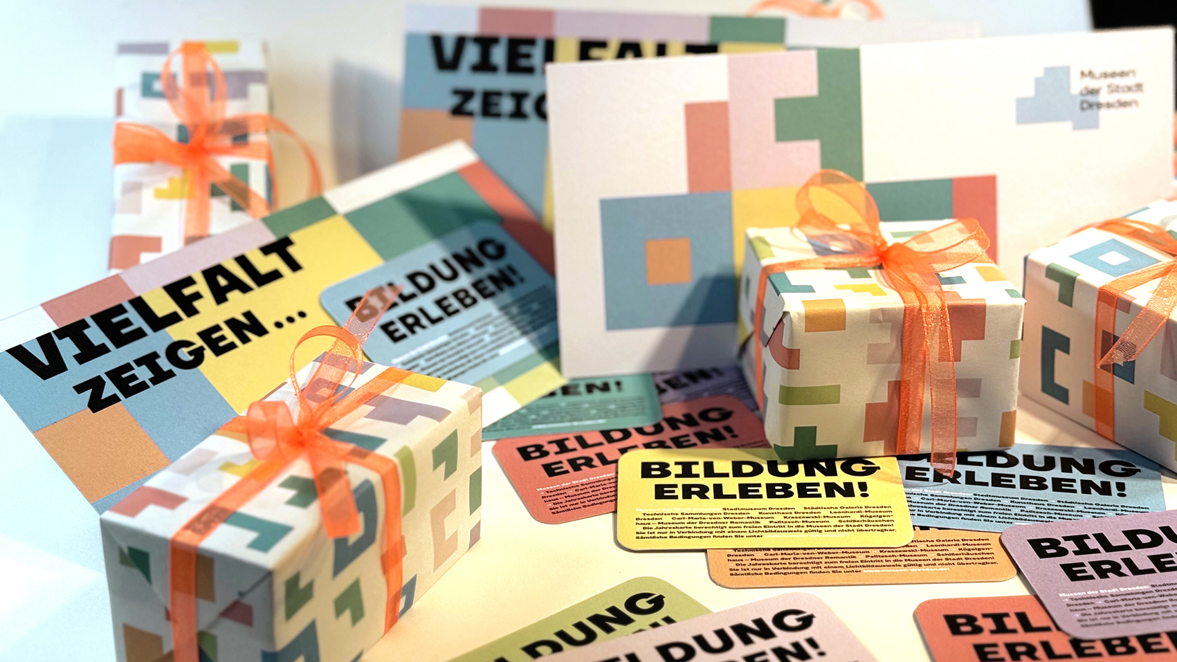 KI generiert: Das Bild zeigt verschiedene bunt verpackte Geschenke mit Schleifen sowie Karten und Flyer mit den Aufschriften "Vielfalt zeigen" und "Bildung erleben!" Es vermittelt eine Botschaft über kulturelle Vielfalt und Bildung, möglicherweise in Zusammenhang mit einem Museum oder einer Bildungseinrichtung.