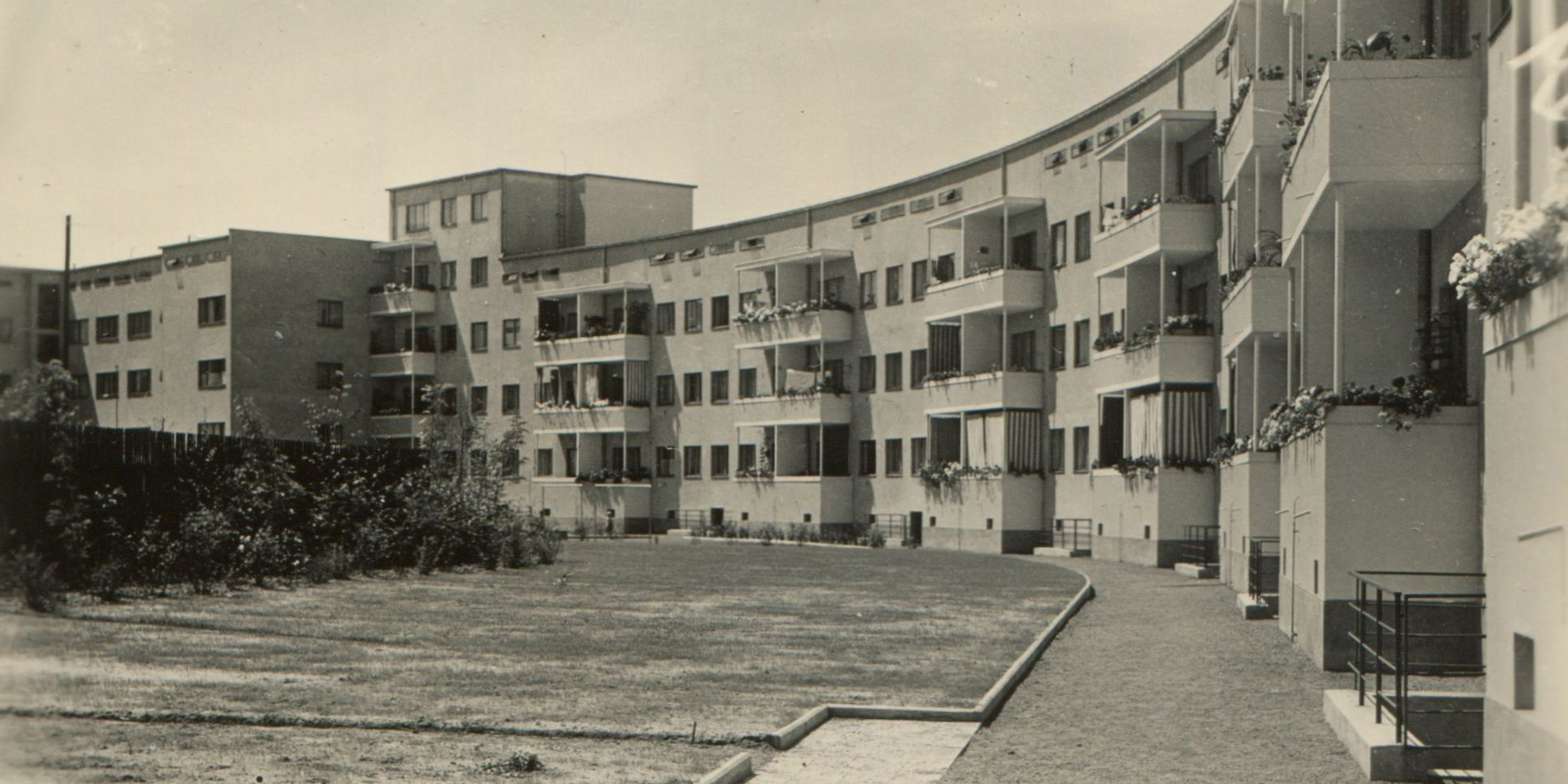 KI generiert: Das Bild zeigt eine Reihe von Wohngebäuden im Stil der klassischen Moderne, die durch ihre sanfte Kurvatur und geometrische Linien auffallen. Im Vordergrund befindet sich eine Grünfläche mit einem Weg, der entlang der Gebäude führt.
