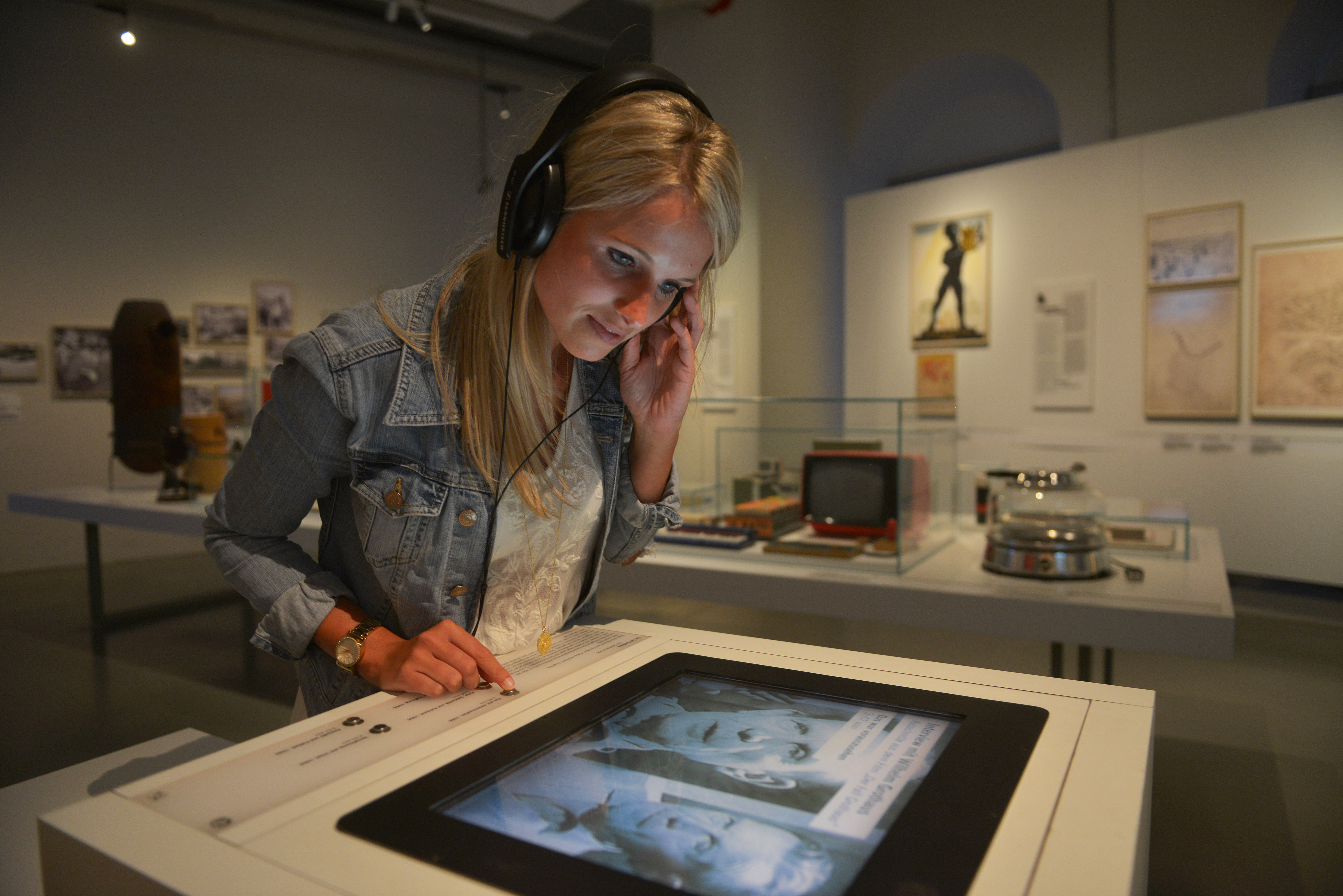 KI generiert: Das Bild zeigt eine Frau, die Kopfhörer trägt und auf einen Bildschirm in einem Museum schaut. Der Hintergrund zeigt verschiedene Ausstellungsstücke und Fotos.