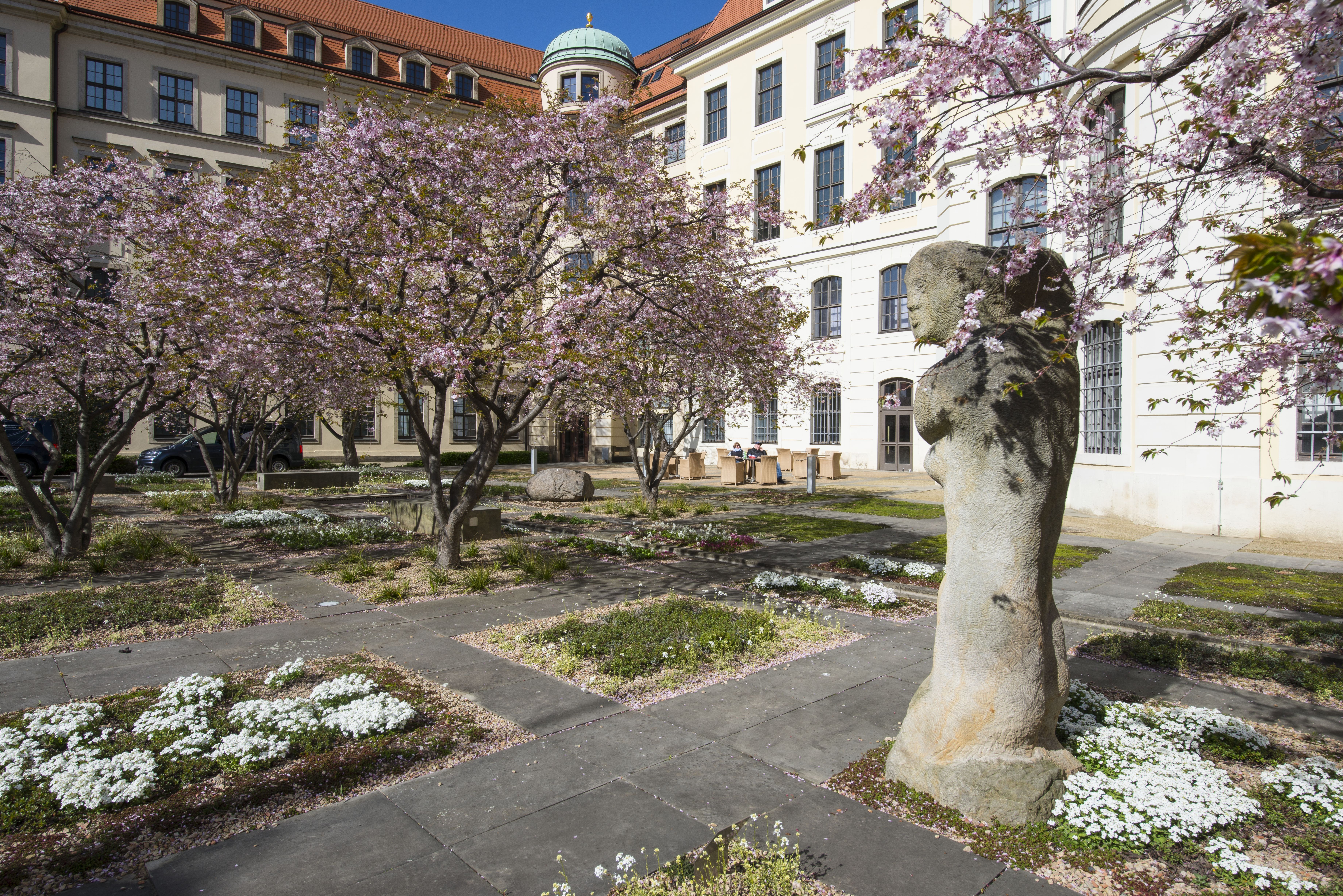 KI generiert: Das Bild zeigt einen Innenhof mit blühenden Bäumen, einem Steingebäude im Hintergrund und einer Skulptur im Vordergrund. Es ist Frühling, und die Blumenbeete sind ebenfalls in voller Blüte.