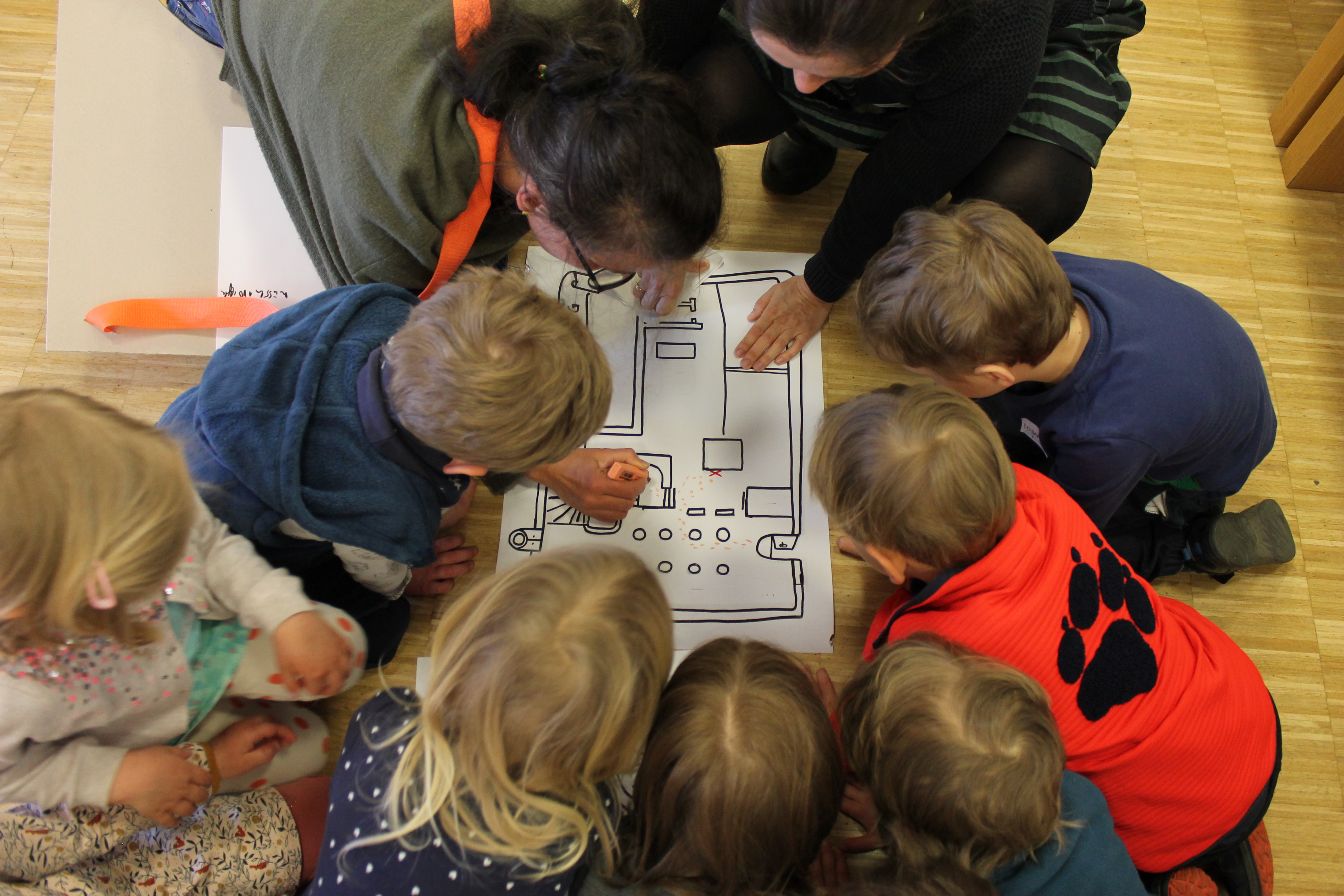 AI generated: Several children and two adults sit around a large plan or diagram and discuss it together. It appears to be a group activity or teaching situation.