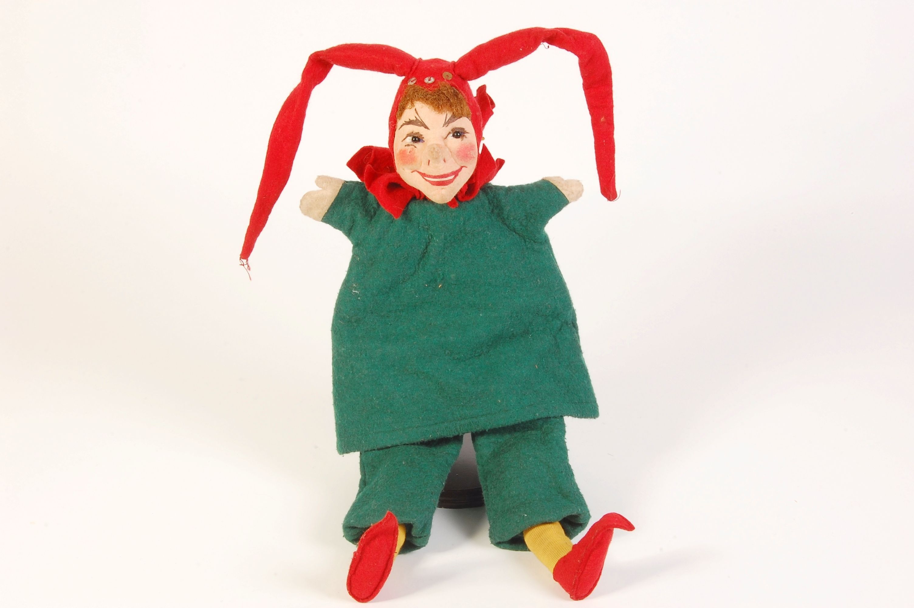 KI generiert: Das Bild zeigt eine Puppe, die ein grünes Kostüm mit roten Schuhen und einer roten Kopfbedeckung trägt. Die Puppe hat ein fröhliches Gesicht und gekreuzte Beine.