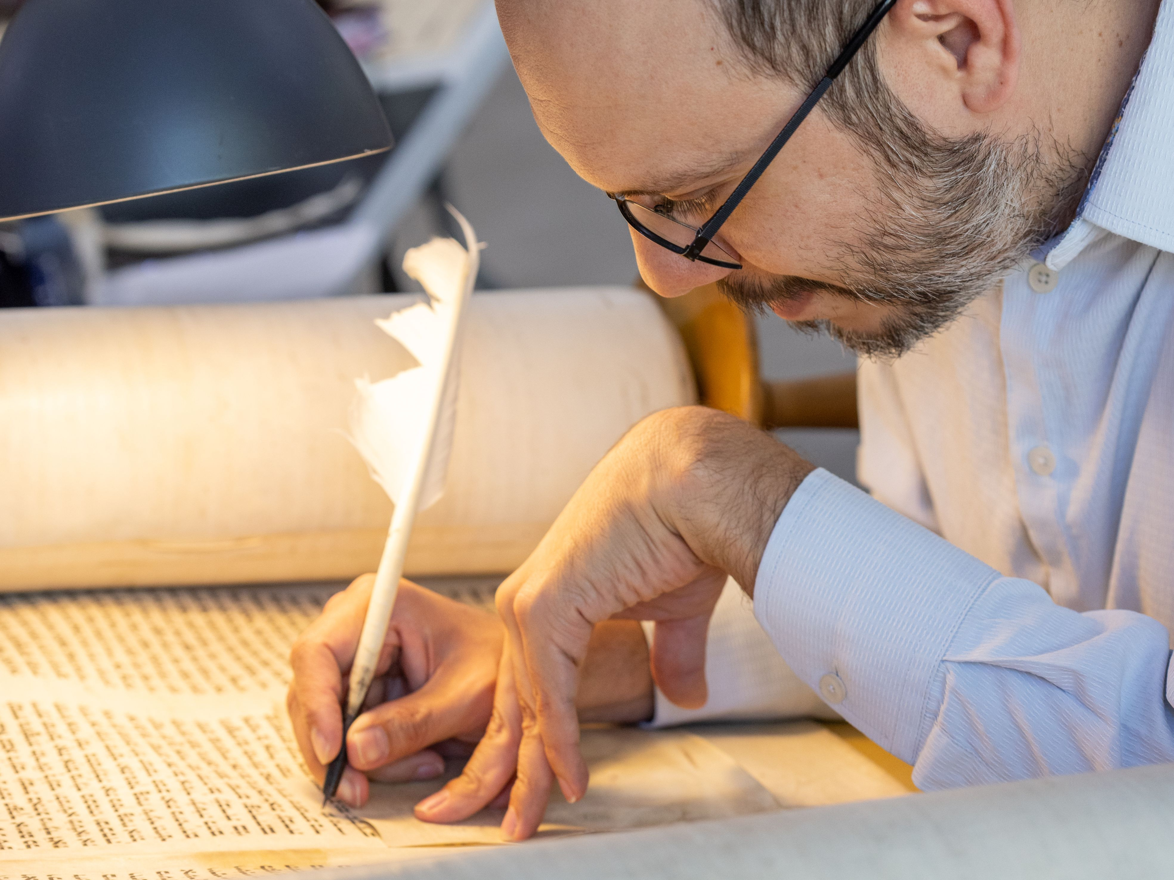 KI generiert: Das Bild zeigt einen Mann, der konzentriert mit einer Feder auf einer Schriftrolle schreibt. Er trägt eine Kippa und arbeitet unter einer Tischlampe.