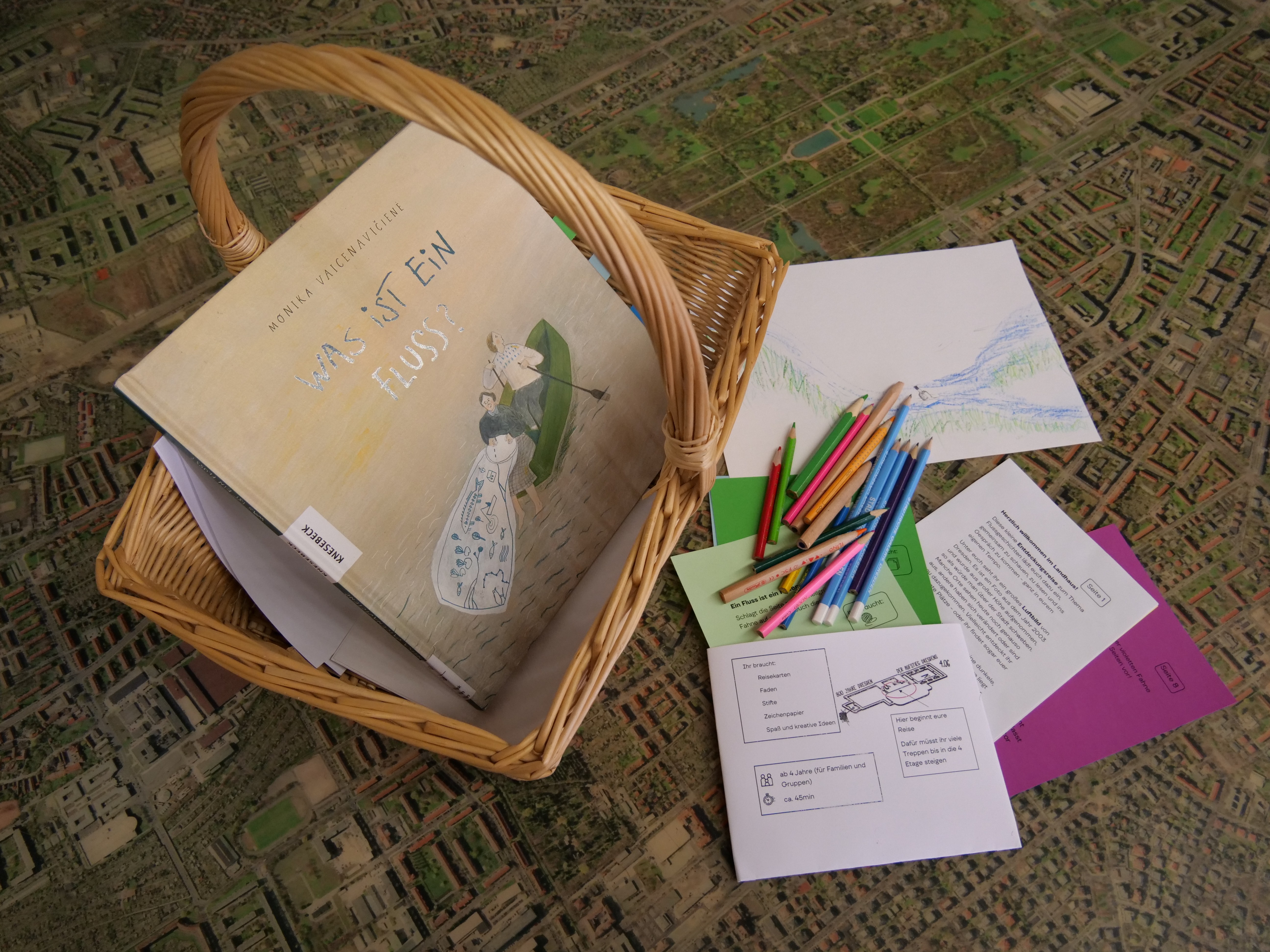 KI generiert: Das Bild zeigt einen geflochtenen Korb mit einem Buch mit dem Titel "Was ist ein Fluss?" und mehreren bunten Buntstiften auf einem Tisch, der eine Art Landkarte oder Stadtplan als Untergrund hat. Neben dem Buch und den Stiften liegen farbige Blätter mit verschiedenen Skizzen und Texten.