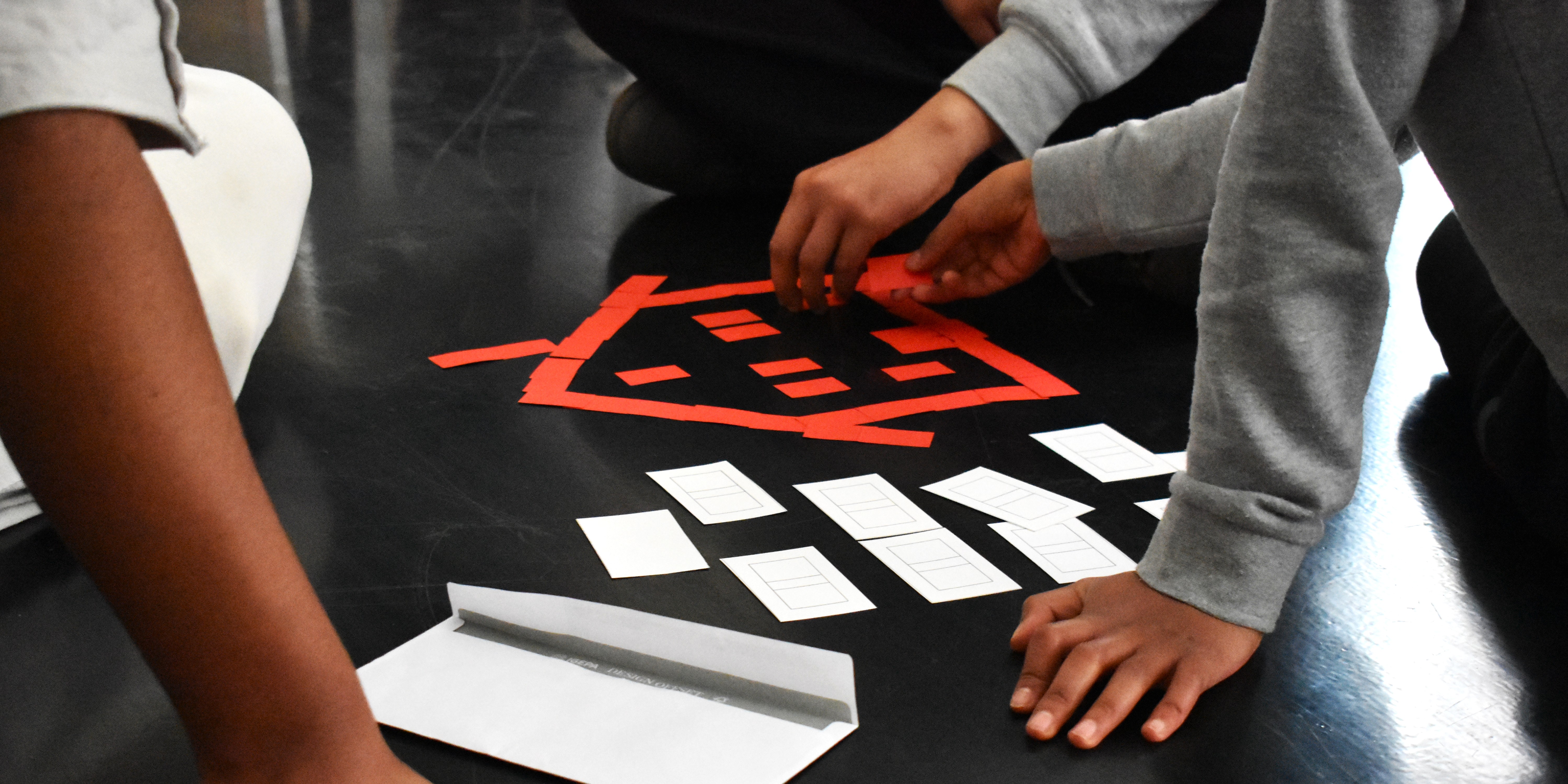 KI generiert: Das Bild zeigt Kinder, die auf dem Boden sitzend mit Karten und roten Klebebändern eine Aktivität durchführen. Sie scheinen Karten oder Papierstücke zu ordnen oder ein Spiel zu spielen.