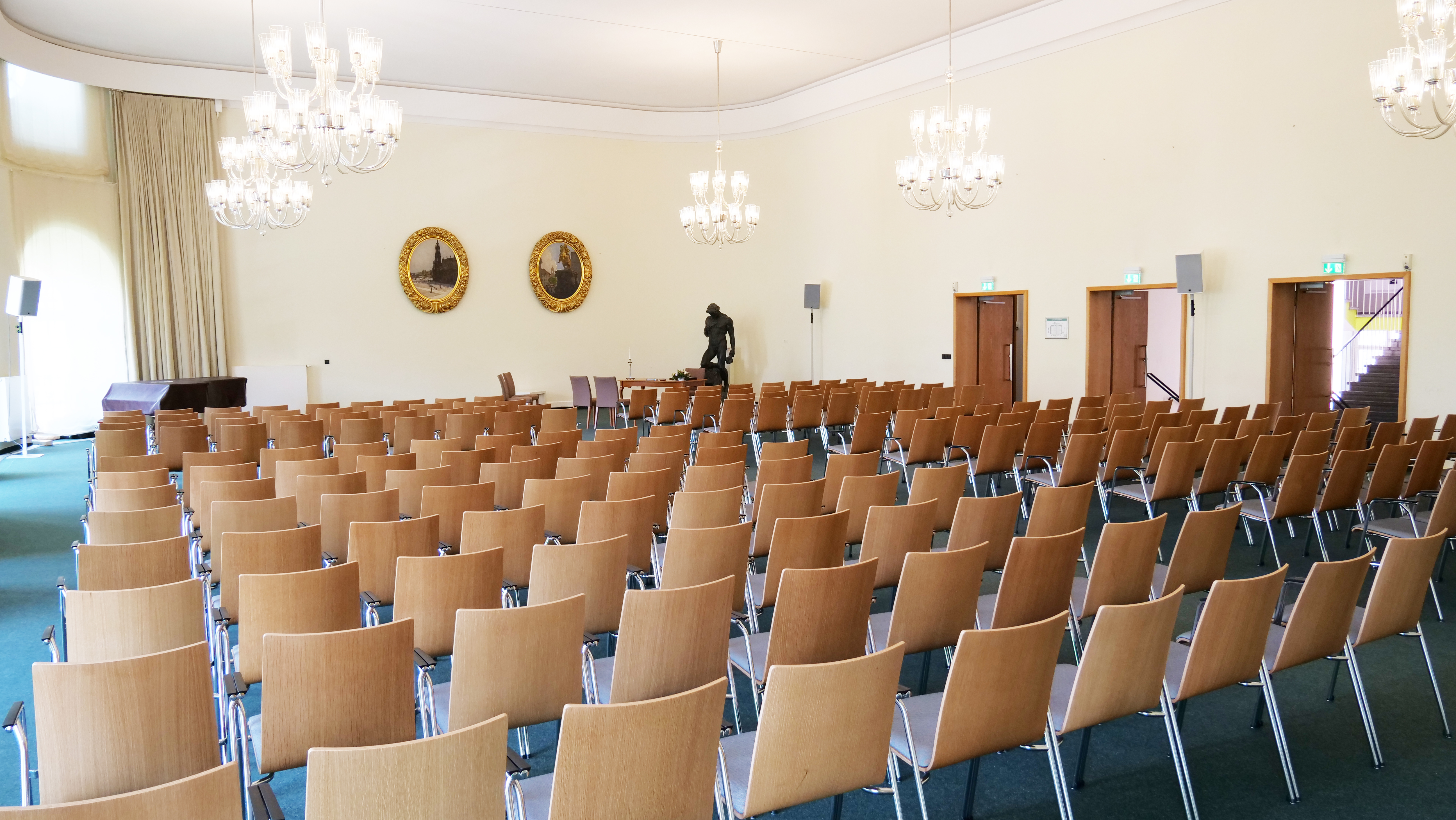 KI generiert: Das Bild zeigt einen leeren Konferenz- oder Vortragssaal, ausgestattet mit mehreren Reihen hölzerner Stühle, Kronleuchtern an der Decke und einer Statue an der hinteren Wand. Im Hintergrund befinden sich Türen, Lautsprecher und zwei große runde Wanddekorationen.