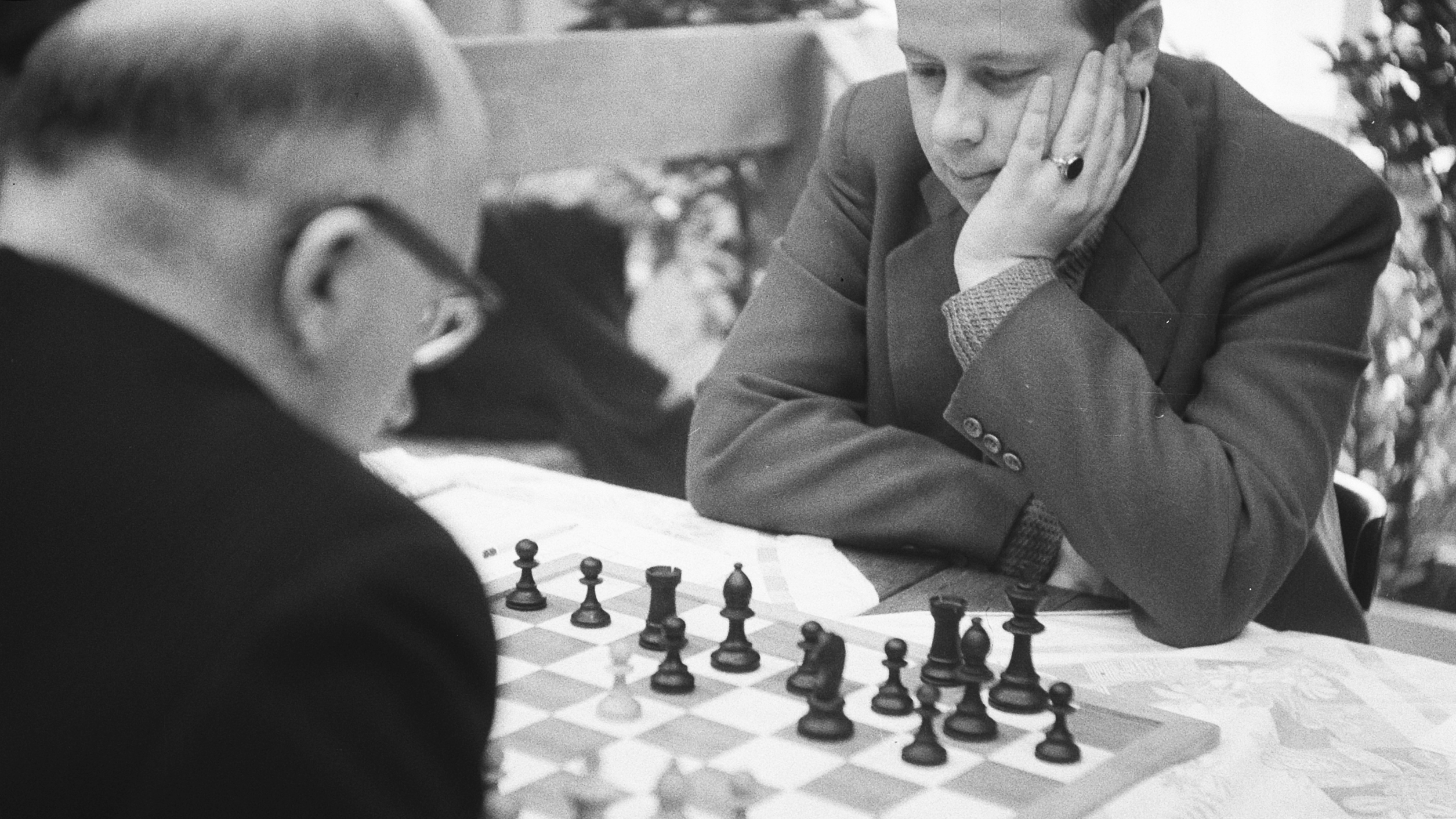 KI generiert: Das Bild zeigt zwei Personen, die konzentriert Schach spielen. Eine Person ist im Vordergrund von hinten zu sehen, während die andere über das Schachbrett gebeugt ist.