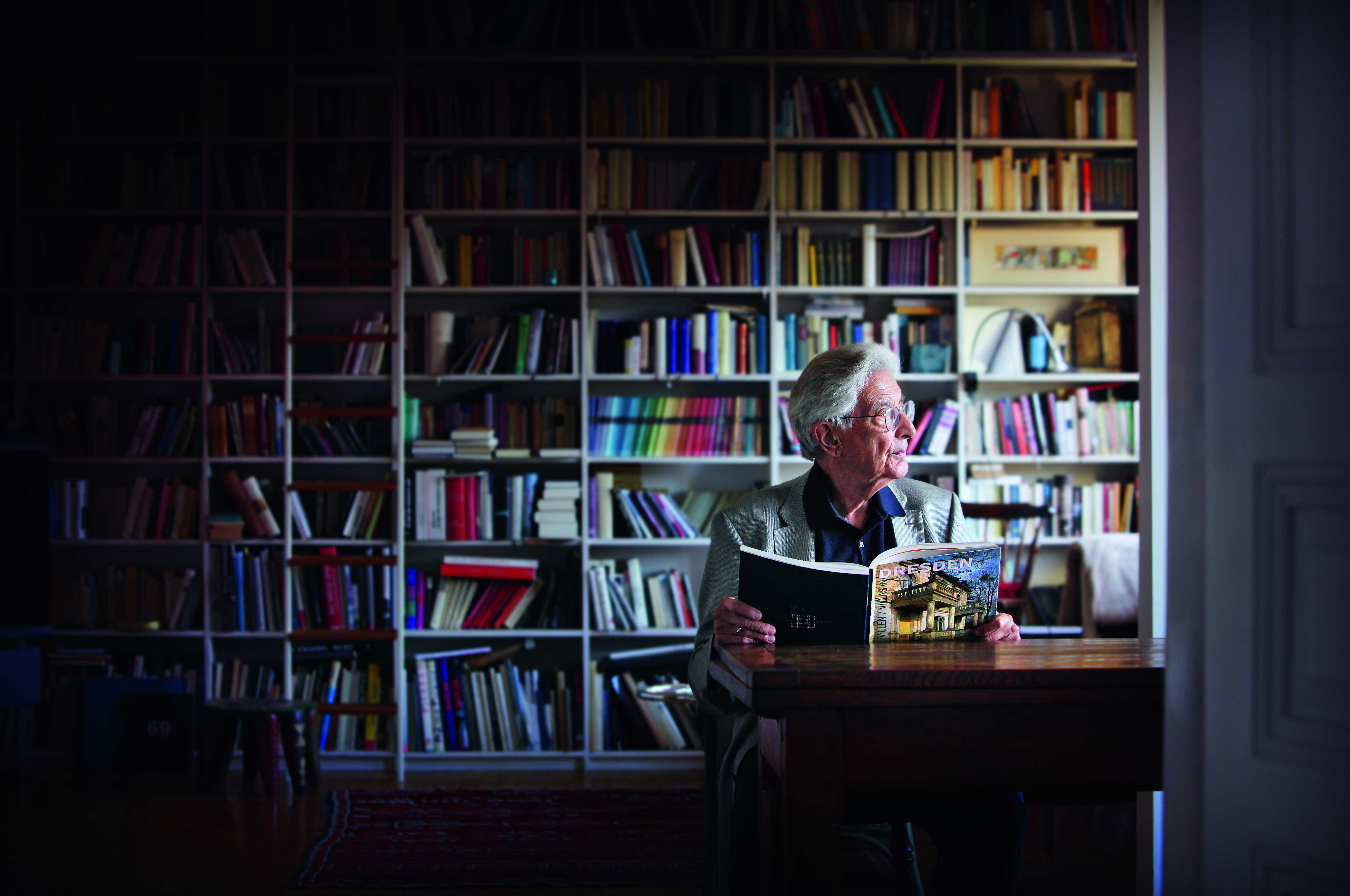KI generiert: Ein älterer Mann sitzt an einem Tisch vor einem großen Bücherregal und hält ein Buch in der Hand. Er scheint nachdenklich zur Seite zu blicken.