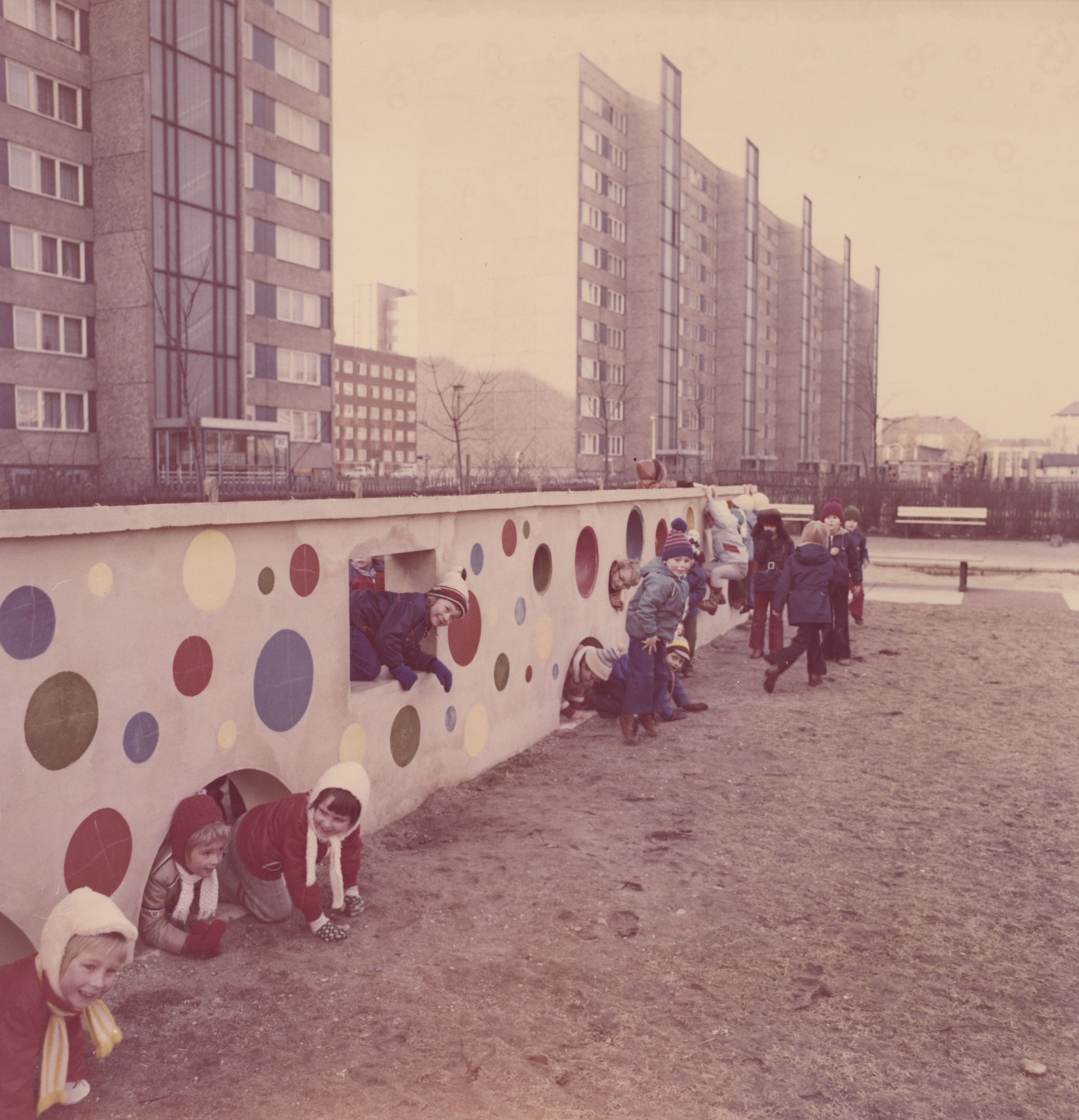 KI generiert: Das Bild zeigt eine Gruppe von Kindern, die auf einem Spielplatz spielen und durch eine bunt bemalte Mauer mit Kreisen krabbeln und klettern. Im Hintergrund sind große Wohngebäude zu sehen.