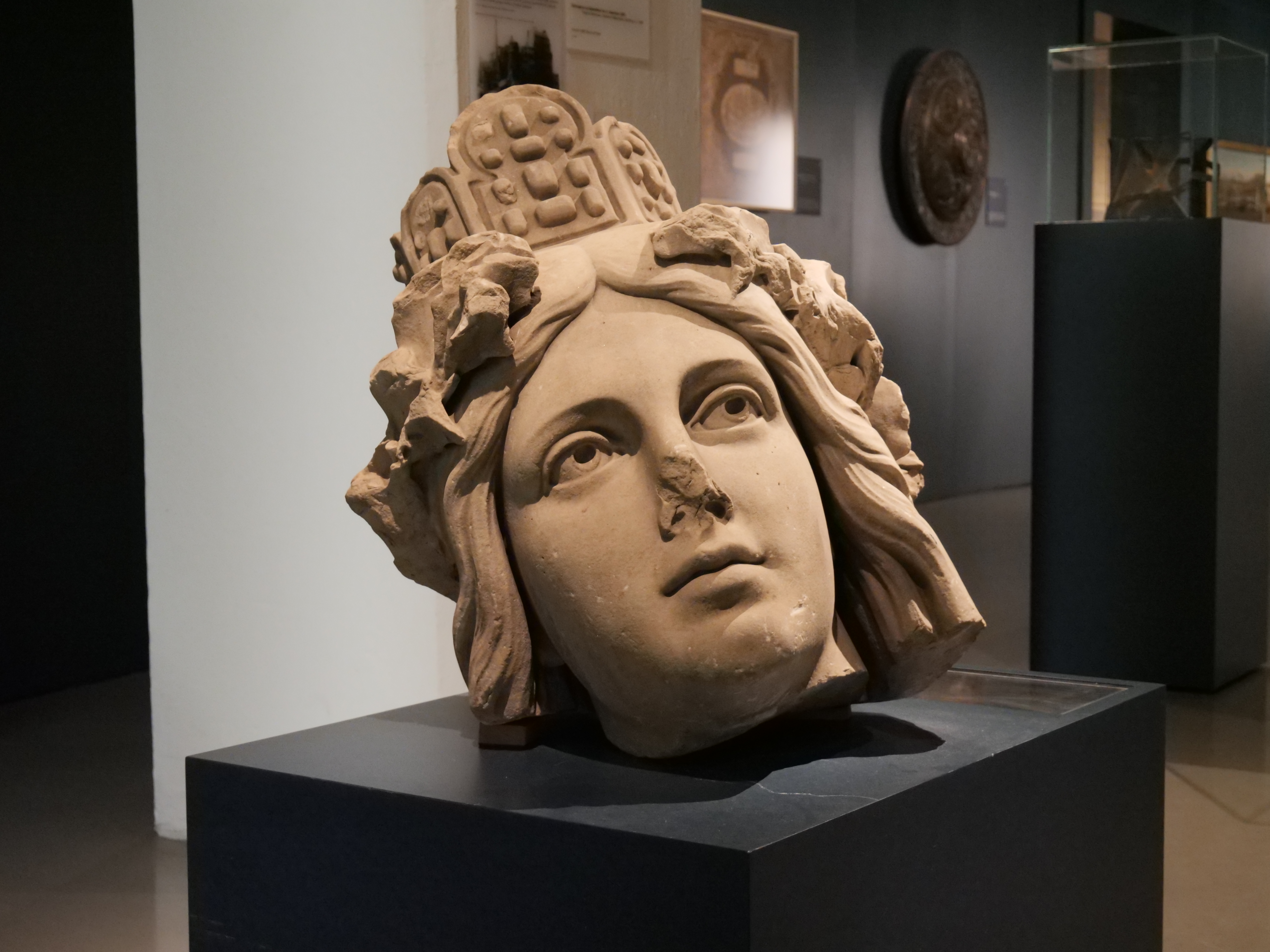 KI generiert: Das Bild zeigt den großen Kopf einer antiken Skulptur, die auf einem Podest in einem Museum ausgestellt ist. Der Kopf ist detailliert gestaltet und weist historische Merkmale wie eine aufwendige Krone oder Kopfschmuck auf.