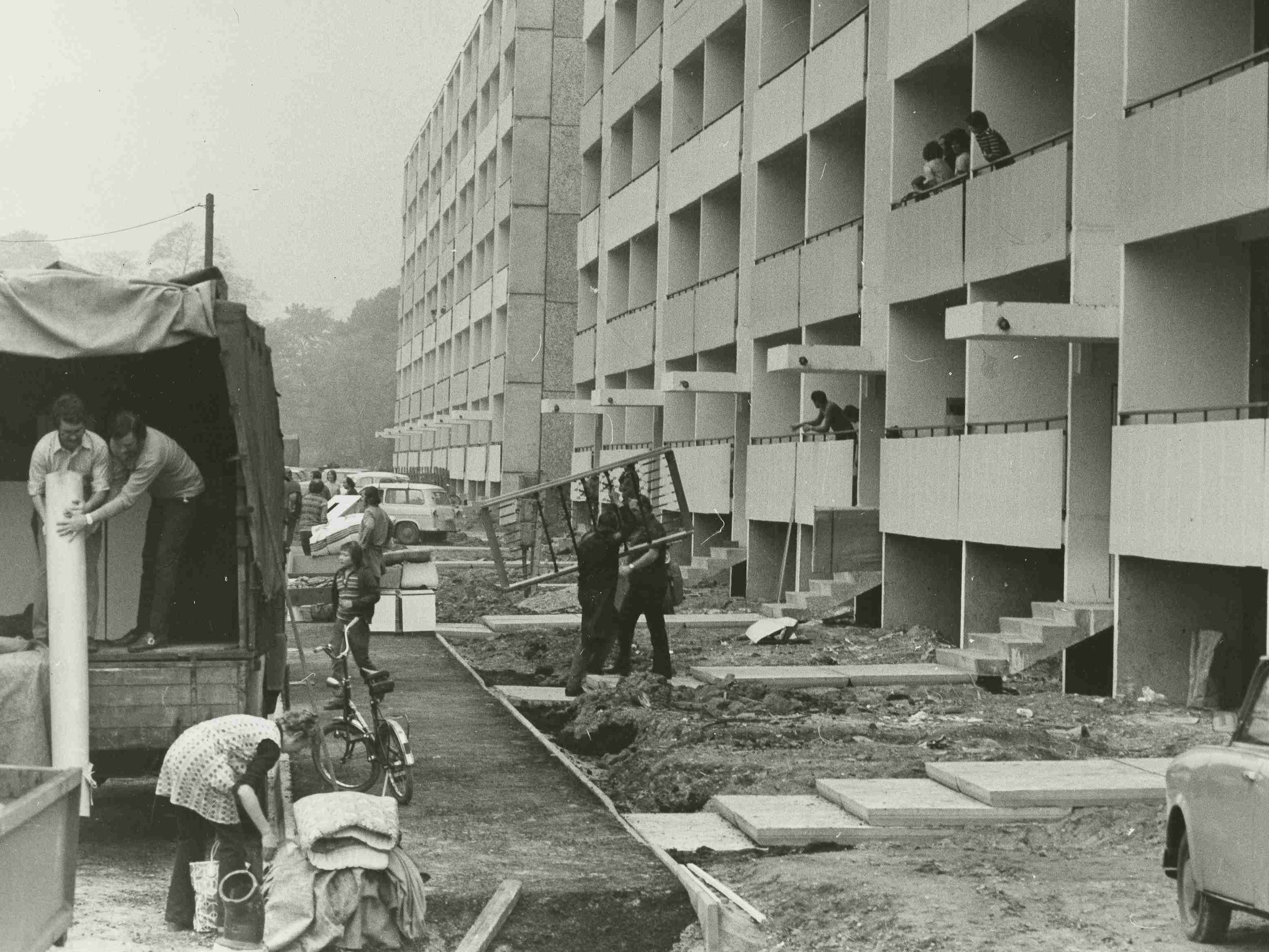 KI generiert: Das Bild zeigt eine Baustelle vor einem mehrstöckigen Wohngebäude, wobei mehrere Personen mit Bauarbeiten und Materiallieferungen beschäftigt sind. Im Vordergrund befinden sich ein LKW, ein Fahrrad und ein Auto, die zur Baustelle gehören.