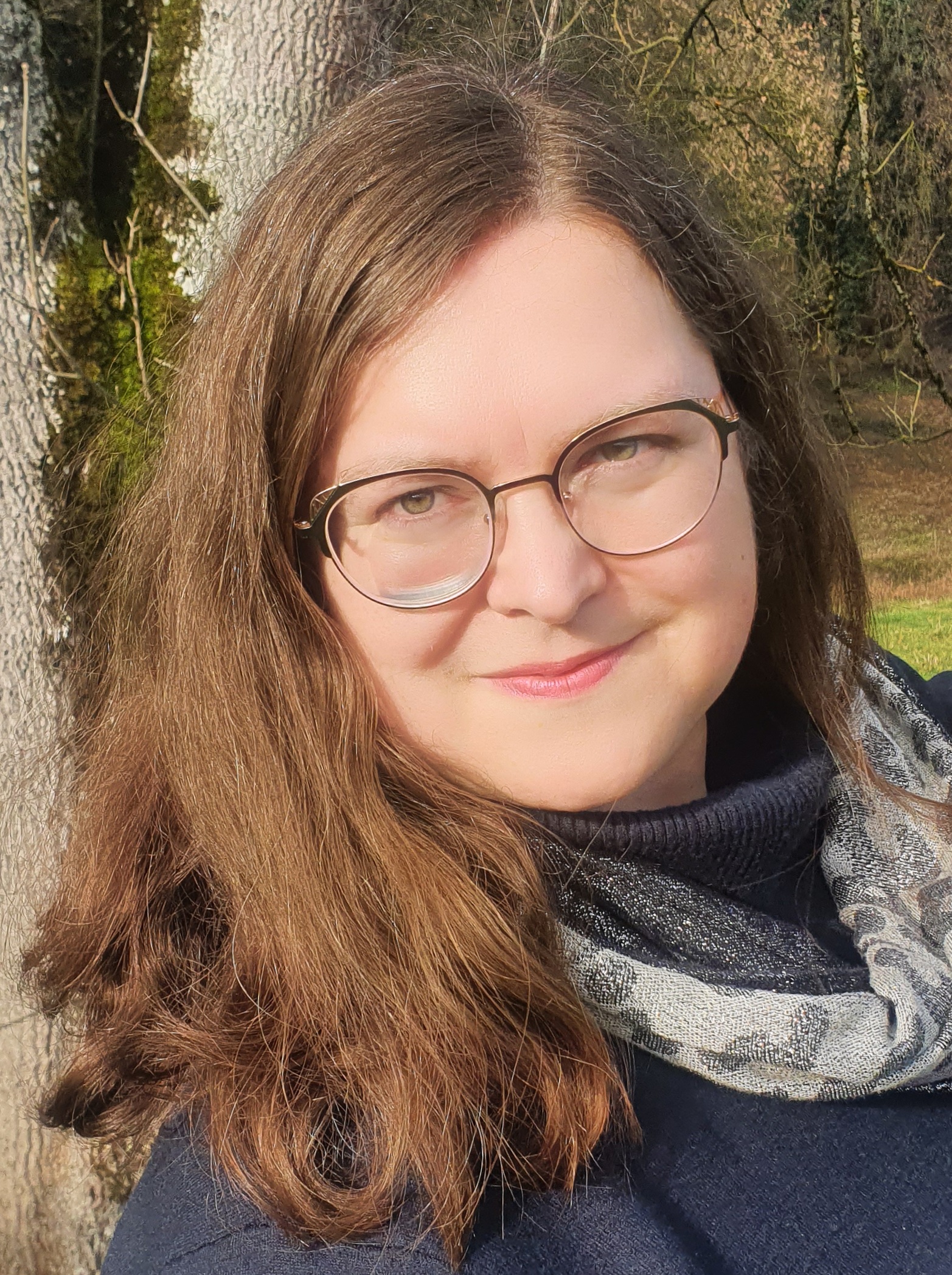 KI generiert: Das Bild zeigt eine Frau mit Brille und braunen Haaren, die draußen vor einem Baum steht. Sie trägt einen Schal und lächelt leicht in die Kamera.