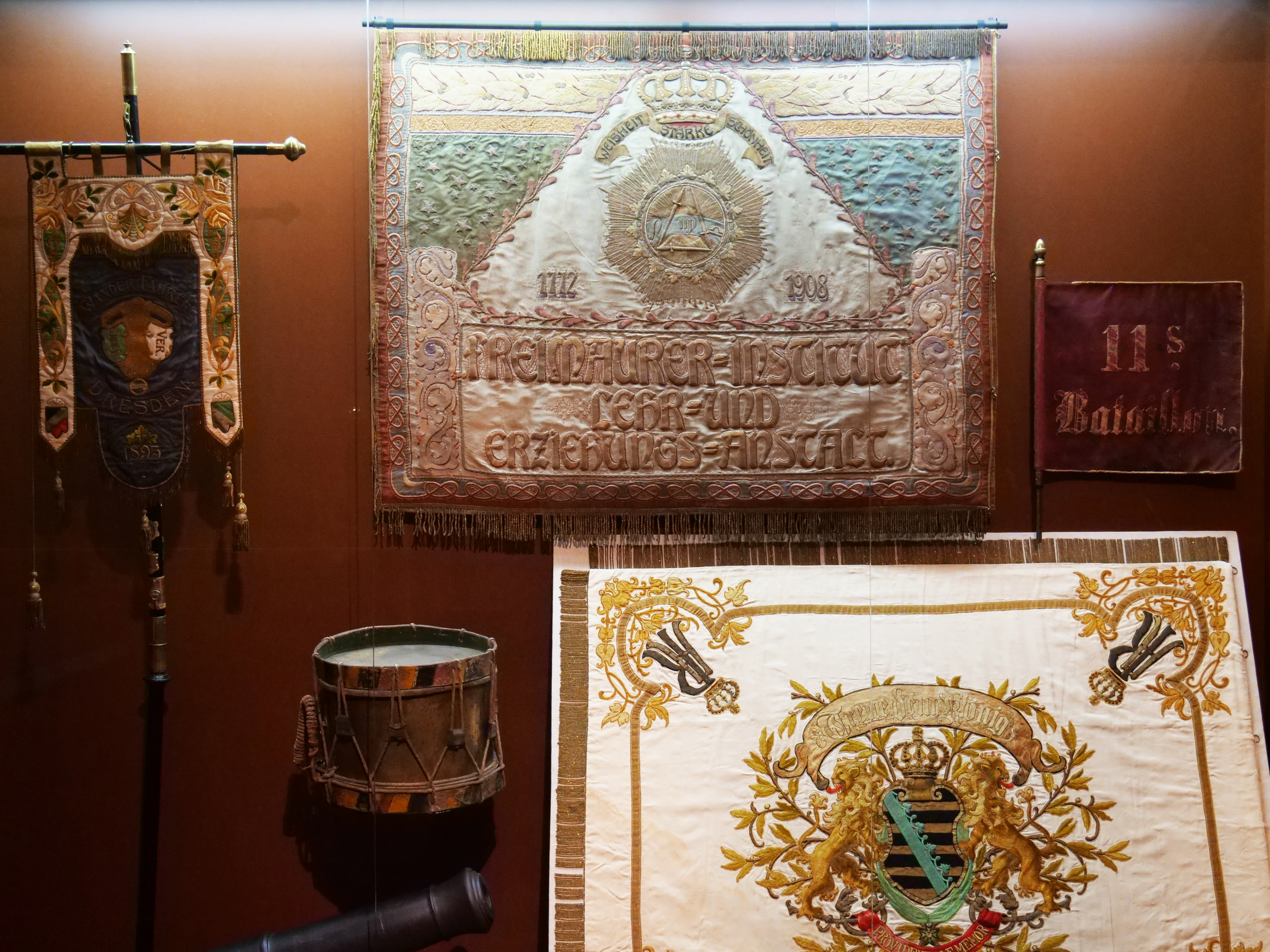 AI generated: The image shows a collection of historical flags and banners that are richly decorated and embroidered with symbols and texts. A drum and a cannon barrel can also be seen in the background.