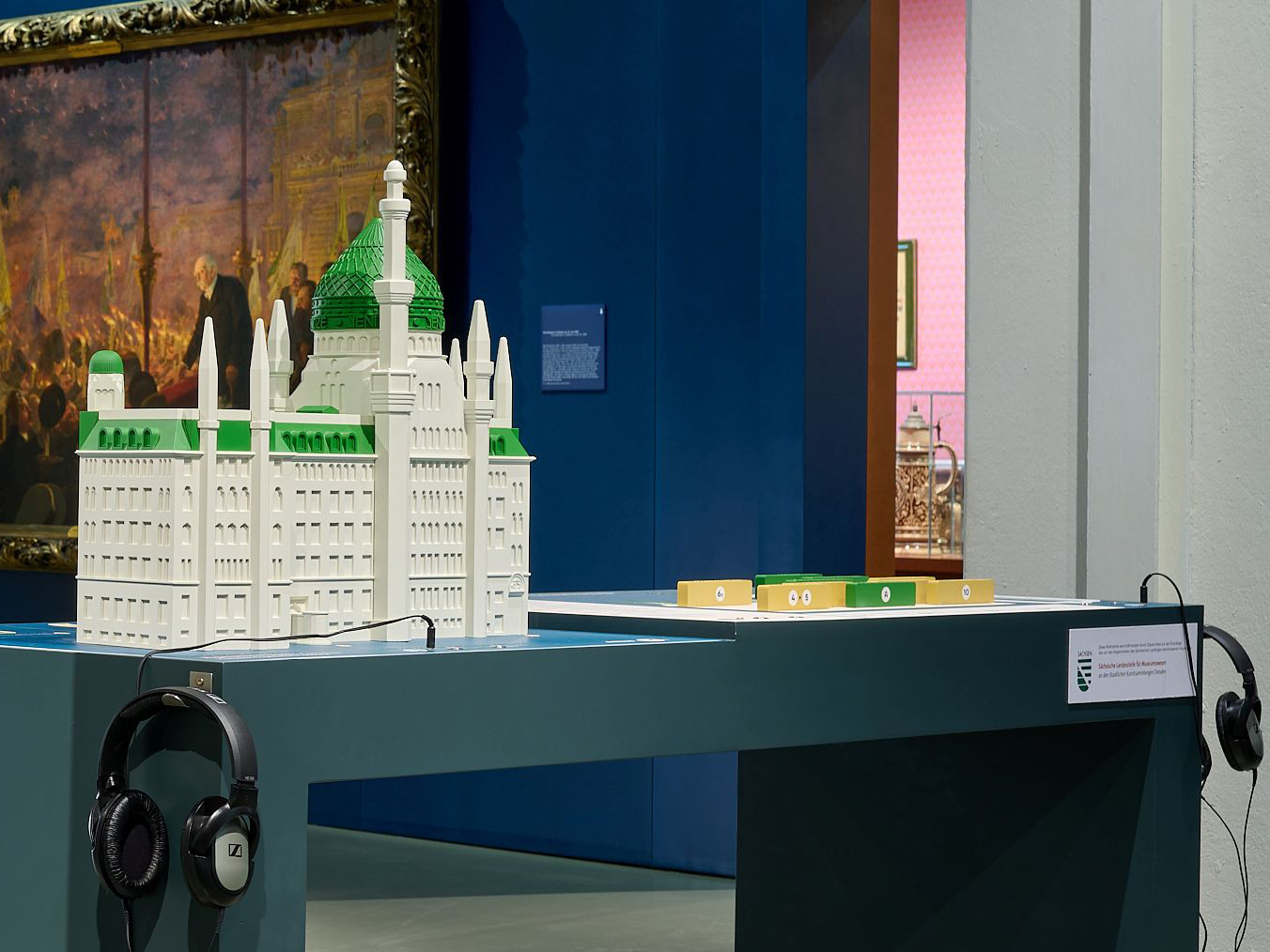 AI-generated: The image shows a model of a building on a blue table, alongside a pair of headphones. A painting can be seen on the wall in the background.