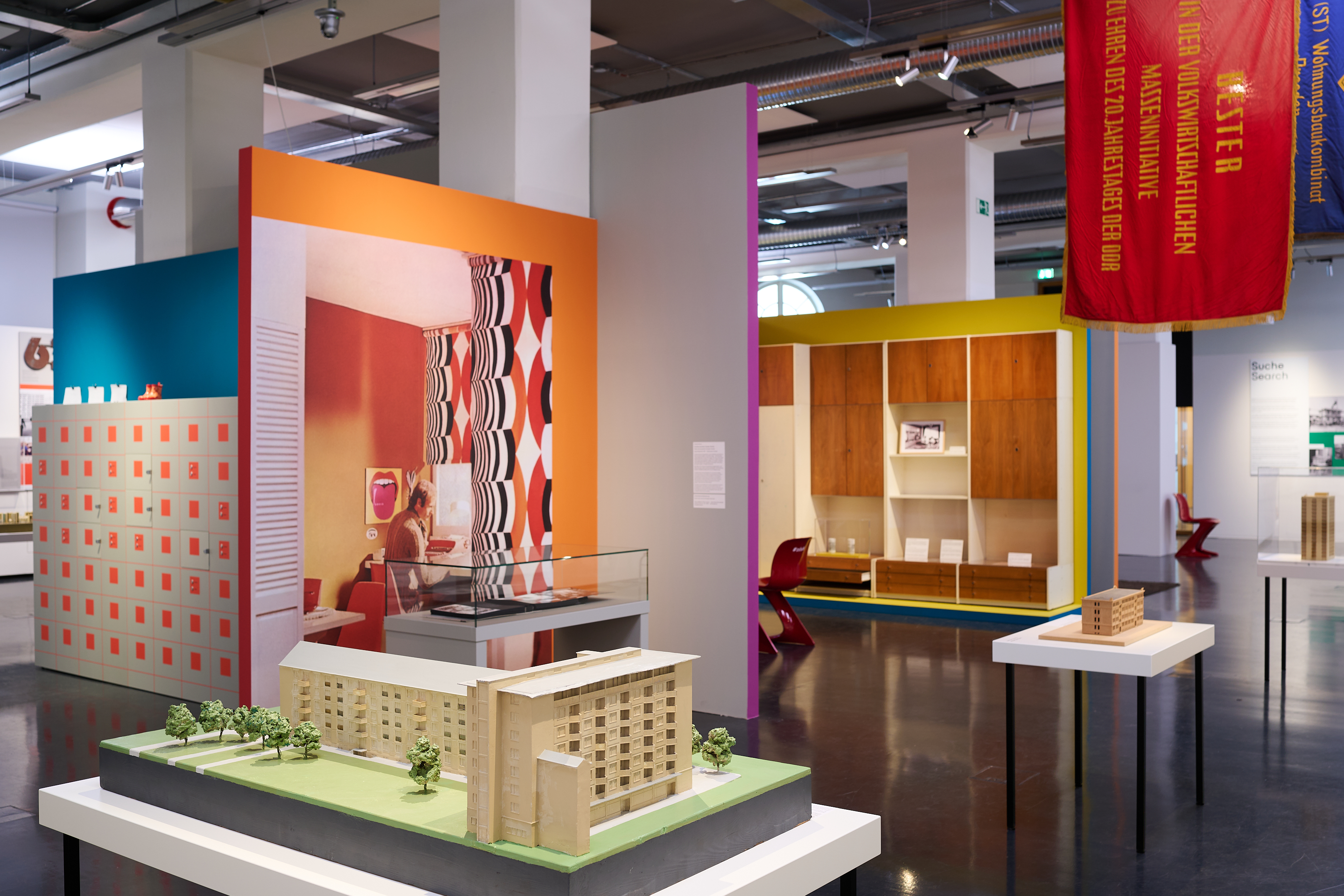 KI generiert: Das Bild zeigt eine Ausstellung mit Architekturmodellen und farbenfrohen Wandelementen, die Innenräume und Gebäude nachbilden. Der Schwerpunkt liegt auf den präsentierten Miniaturgebäuden und der kreativen Gestaltung des Ausstellungsraums.