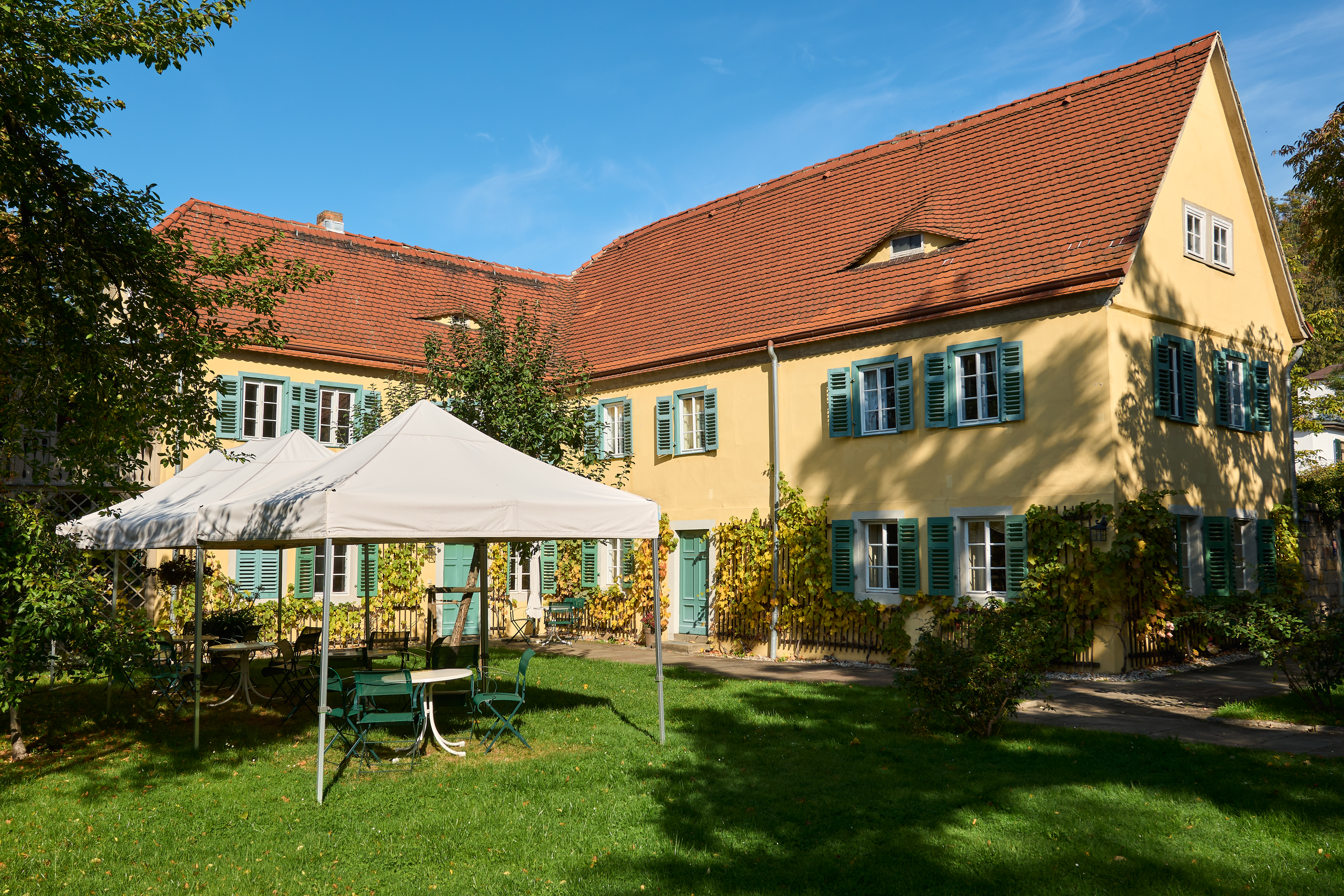 KI generiert: Das Bild zeigt einen gelben, zweistöckigen Gebäudekomplex mit roten Ziegeldächern und grünen Fensterläden in einem Garten mit einem weißen Pavillon und grünen Gartenmöbeln. Der Himmel ist klar und blau, und es ist sonnig.