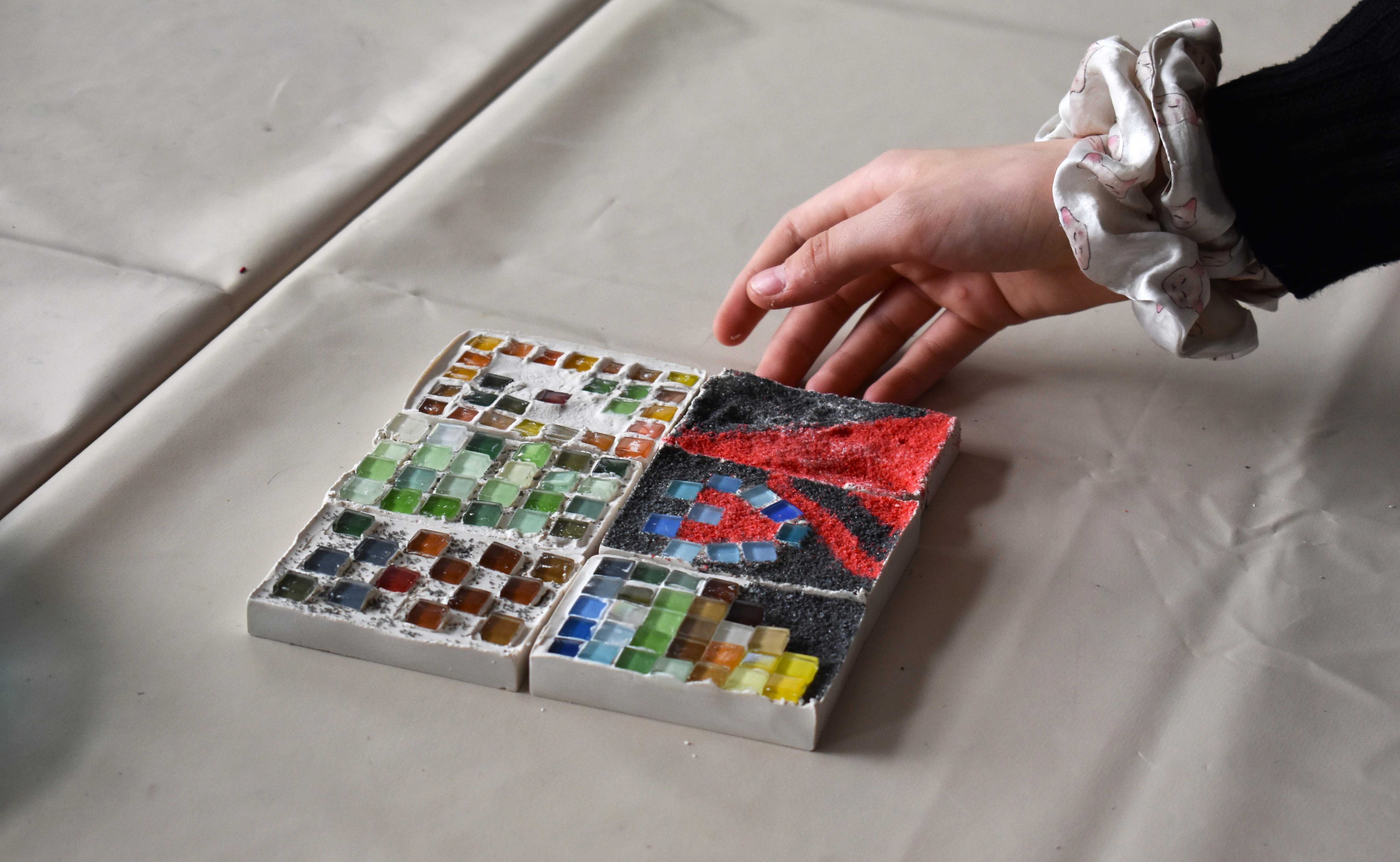 KI generiert: Das Bild zeigt eine Hand, die sich einem Mosaik aus bunten Glassteinen und Sand nähert. Das Mosaik besteht aus vier Quadraten, die verschiedene Muster und Farben aufweisen.