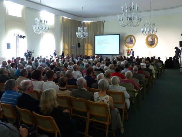 KI generiert: Das Bild zeigt einen vollen Konferenzraum mit vielen sitzenden Zuhörern, die auf eine Leinwand und eine Rednerbühne blicken. Es handelt sich vermutlich um eine Präsentation oder ein Vortrag, da vorne ein Rednerpult und ein Projektorbildschirm zu sehen sind.