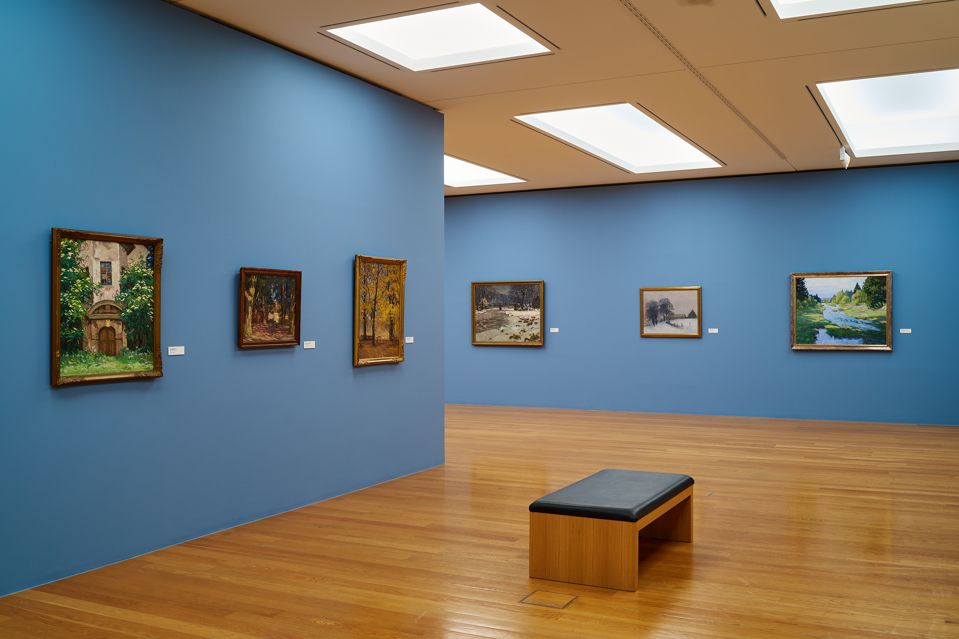 KI generiert: Das Bild zeigt einen Ausstellungsraum in einem Museum mit blauen Wänden, an denen mehrere Gemälde hängen. Im Vordergrund befindet sich eine Bank auf einem Parkettboden.