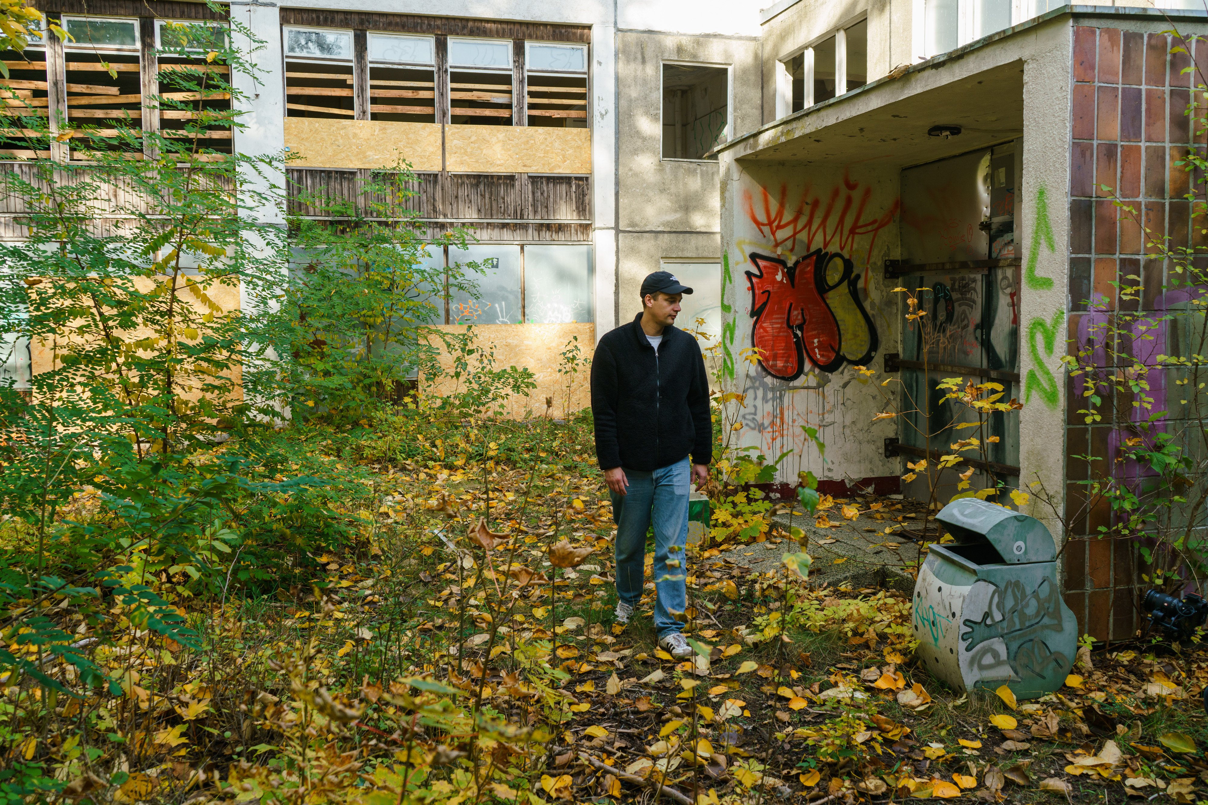 KI generiert: Ein Mann spaziert durch einen verlassenen und überwucherten Bereich mit einem Graffiti-bedeckten Gebäude im Hintergrund. Die Umgebung ist mit Laub bedeckt und wirkt urban-verfallen.