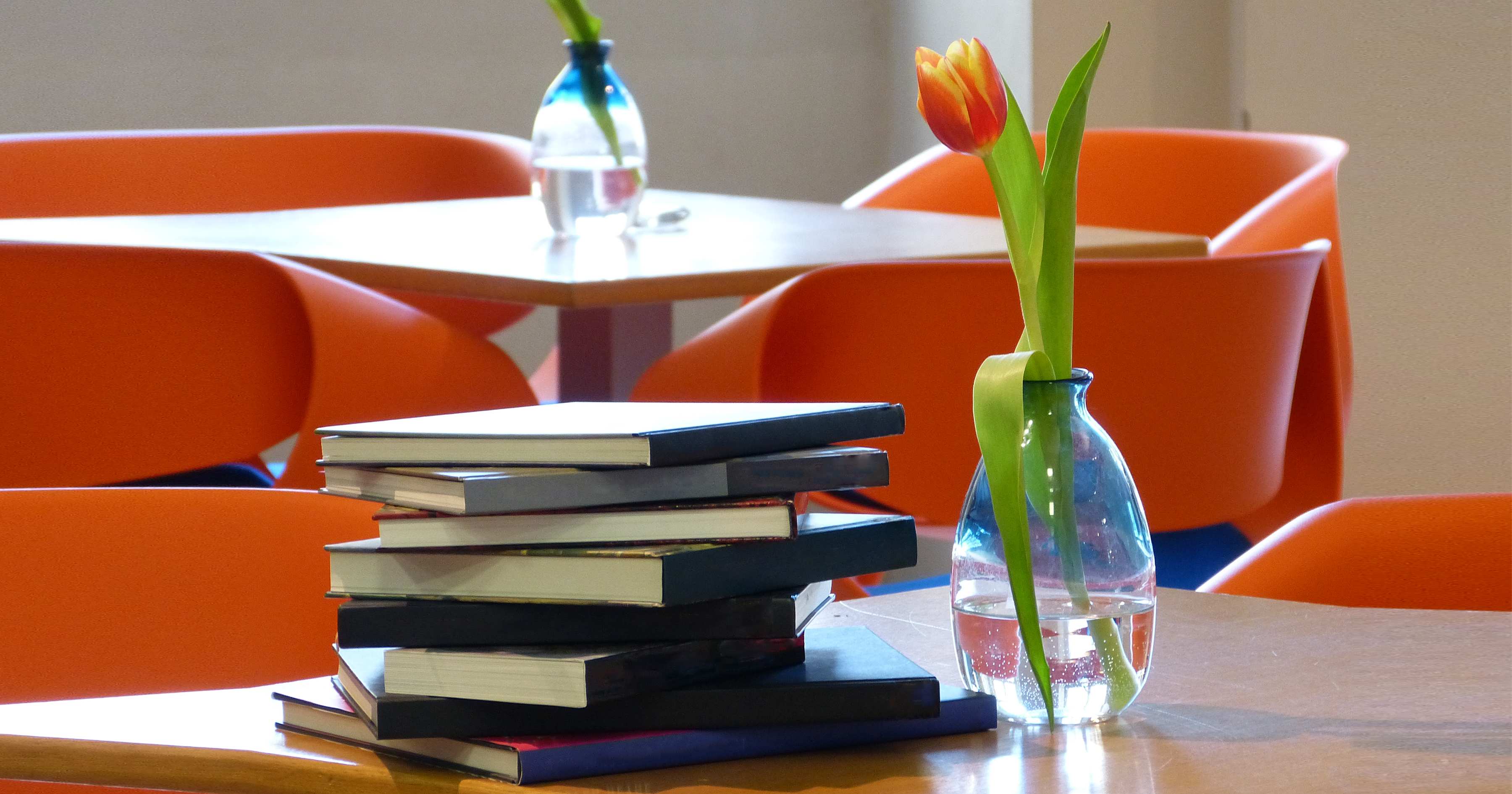 KI generiert: Das Bild zeigt einen Stapel Bücher und eine Vase mit einer einzelnen Tulpe auf einem Tisch, während im Hintergrund mehrere orangefarbene Stühle und weitere Tische zu sehen sind. Der Hauptinhalt des Bildes ist eine ruhige, möglicherweise studienbezogene Atmosphäre in einem modernen Raum.