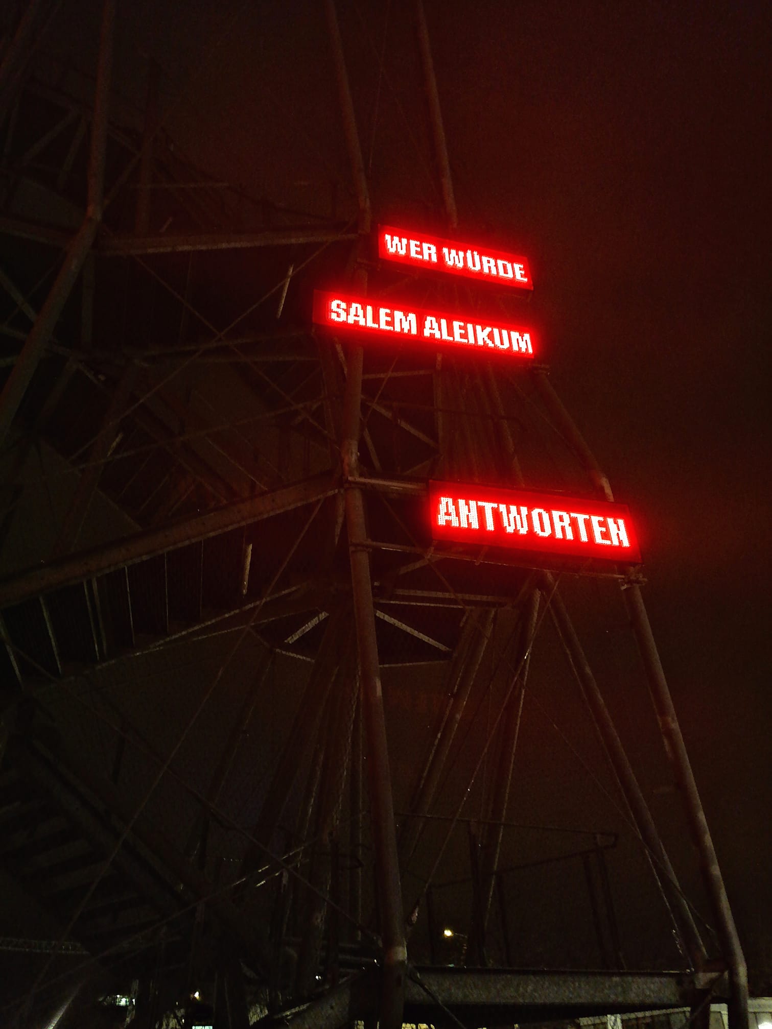 Eine LED-Installation an der Fluchttreppe des Landhauses zeigt aufgeteilt in drei Abschnitte untereinander die Worte "WER WÜRDE / SALEM ALEIKUM / ANTWORTEN" 